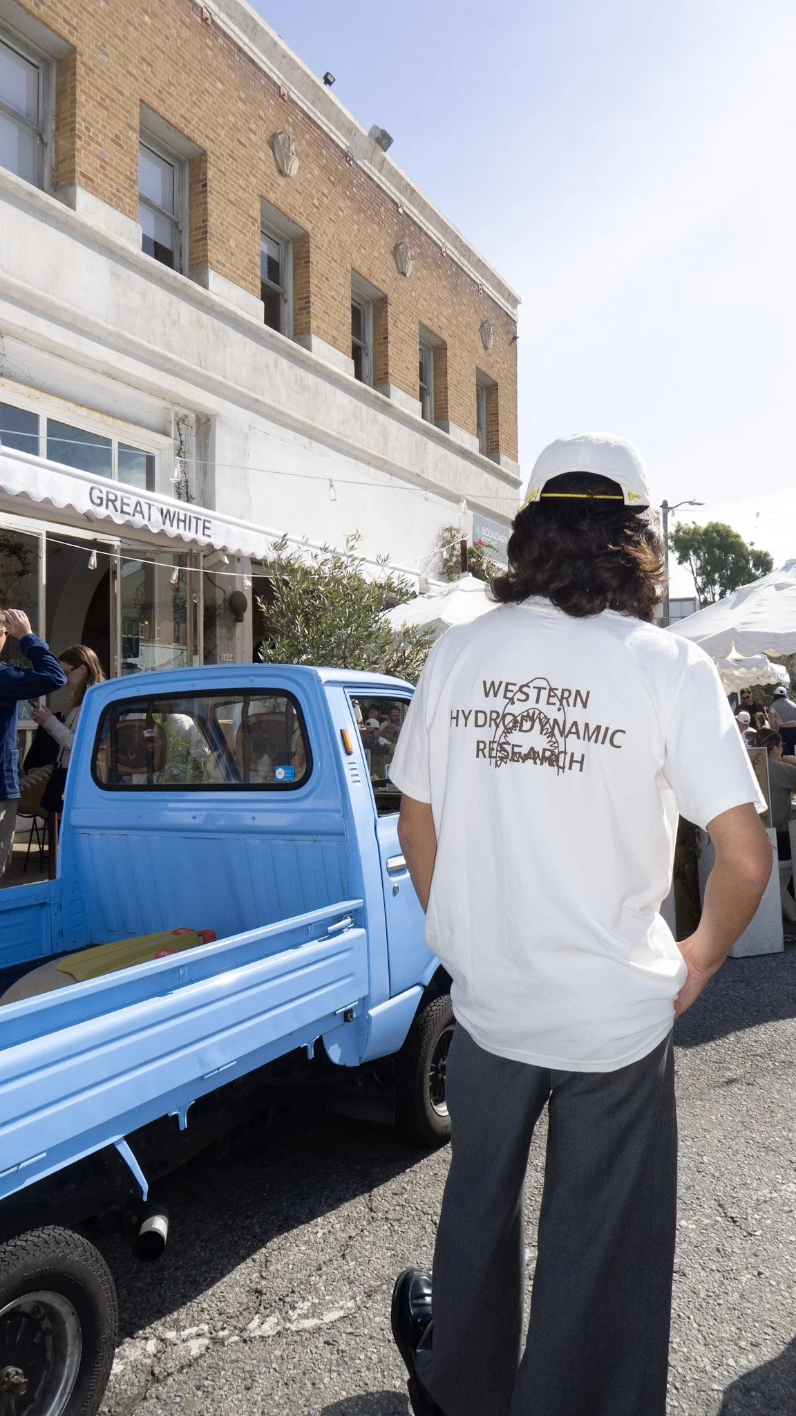 Person wearing a white t-shirt with the words "Western Hydrodynamic Research" and a graphic of a dolphin, standing next to a light blue mini truck at an outdoor event on a sunny day.