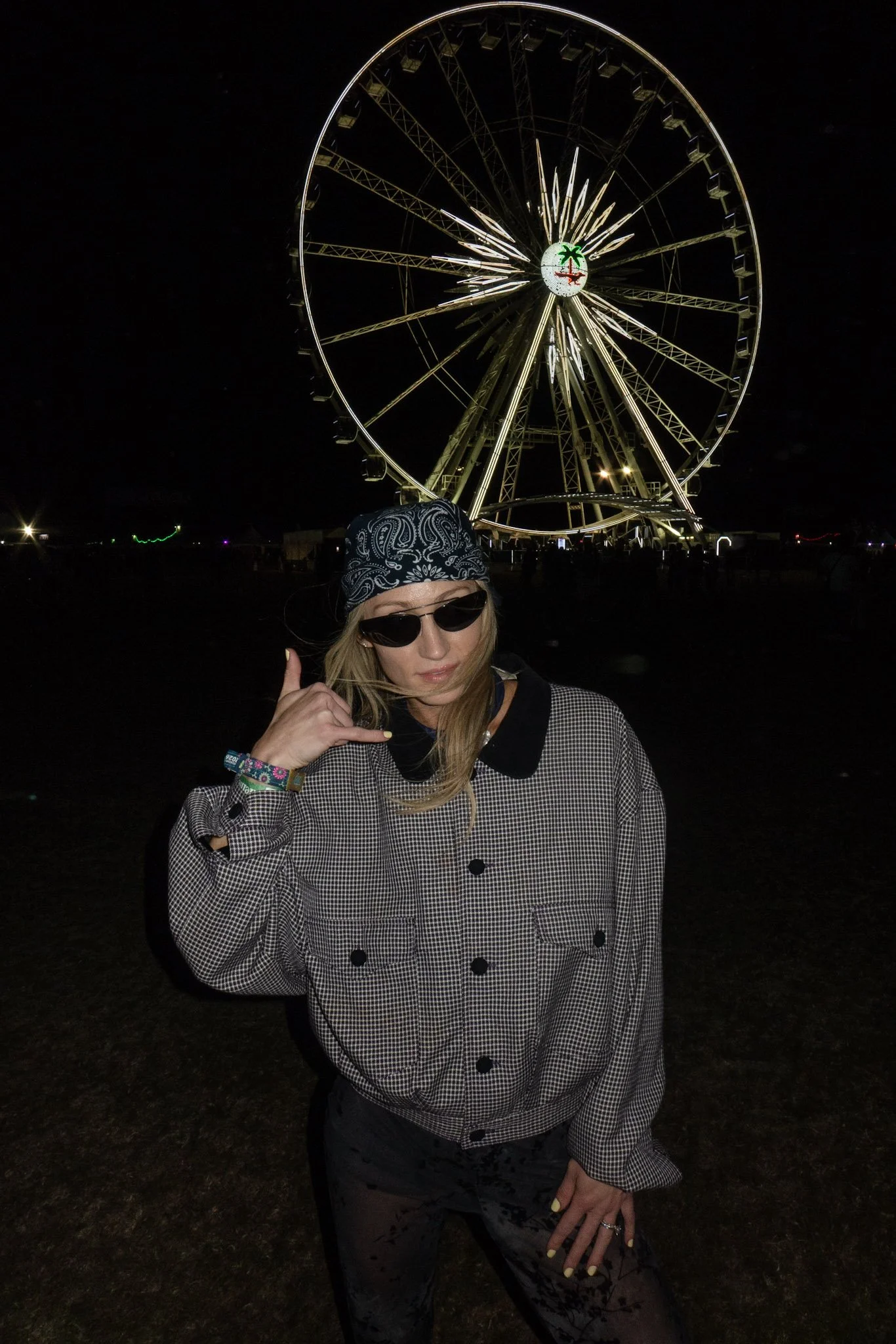 A woman with blonde hair wearing dark sunglasses, a black bandana, and a checkered jacket, posing outdoors at night with a large, illuminated Ferris wheel in the background.