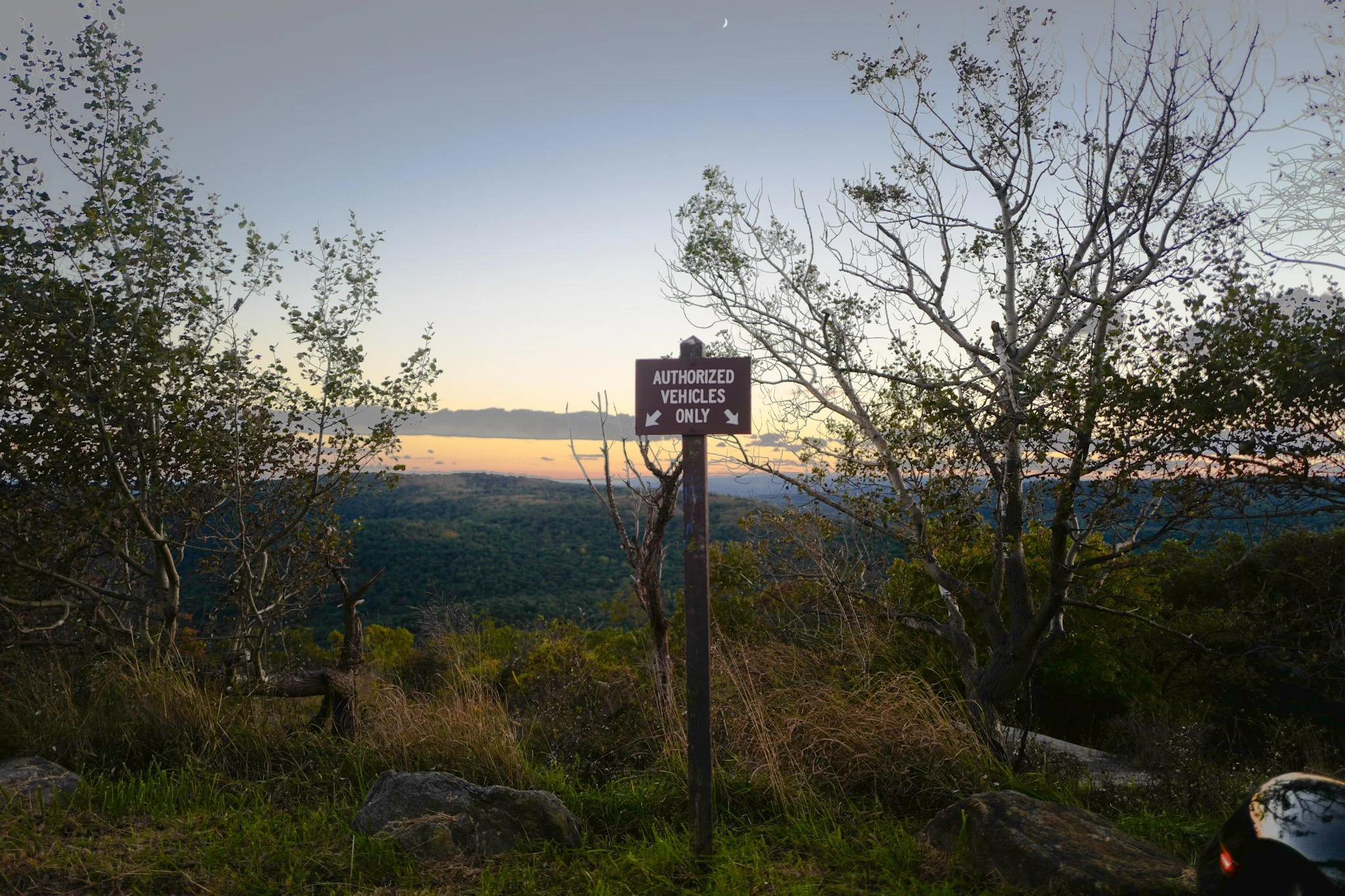 Landscape view at sunset or dusk with a sign that reads 'Authorized Vehicles Only,' trees, hills, and a clear sky with a moon visible.