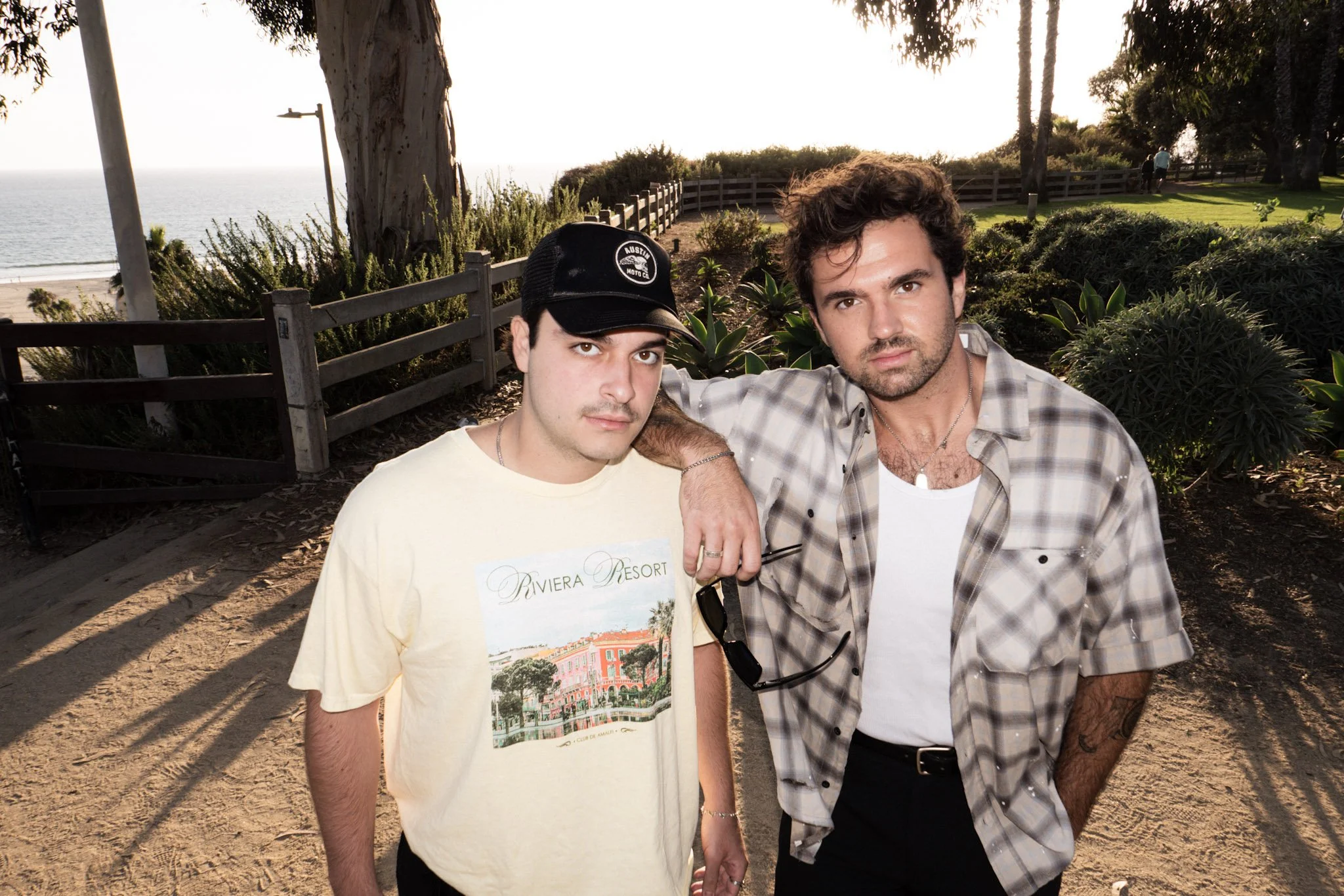 Two young men standing outdoors at a park near the ocean during sunset. One wearing a cap and beige T-shirt with a Riviera Resort print, the other with curly hair in a checkered shirt. They are close together, with the blond man's arm around the othe