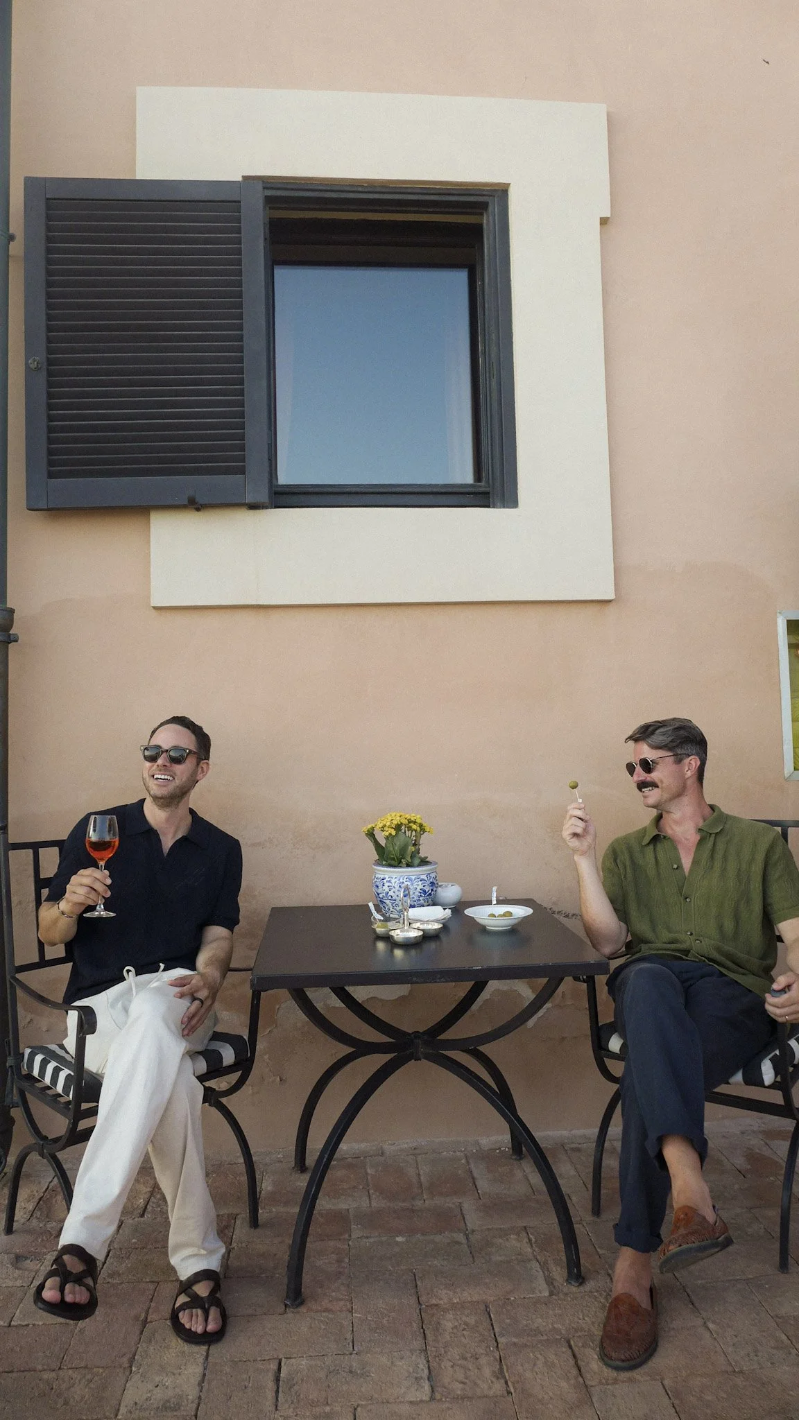Two men sitting at an outdoor table, smiling and holding drinks, with a potted flower on the table, against a peach-colored wall with a window.