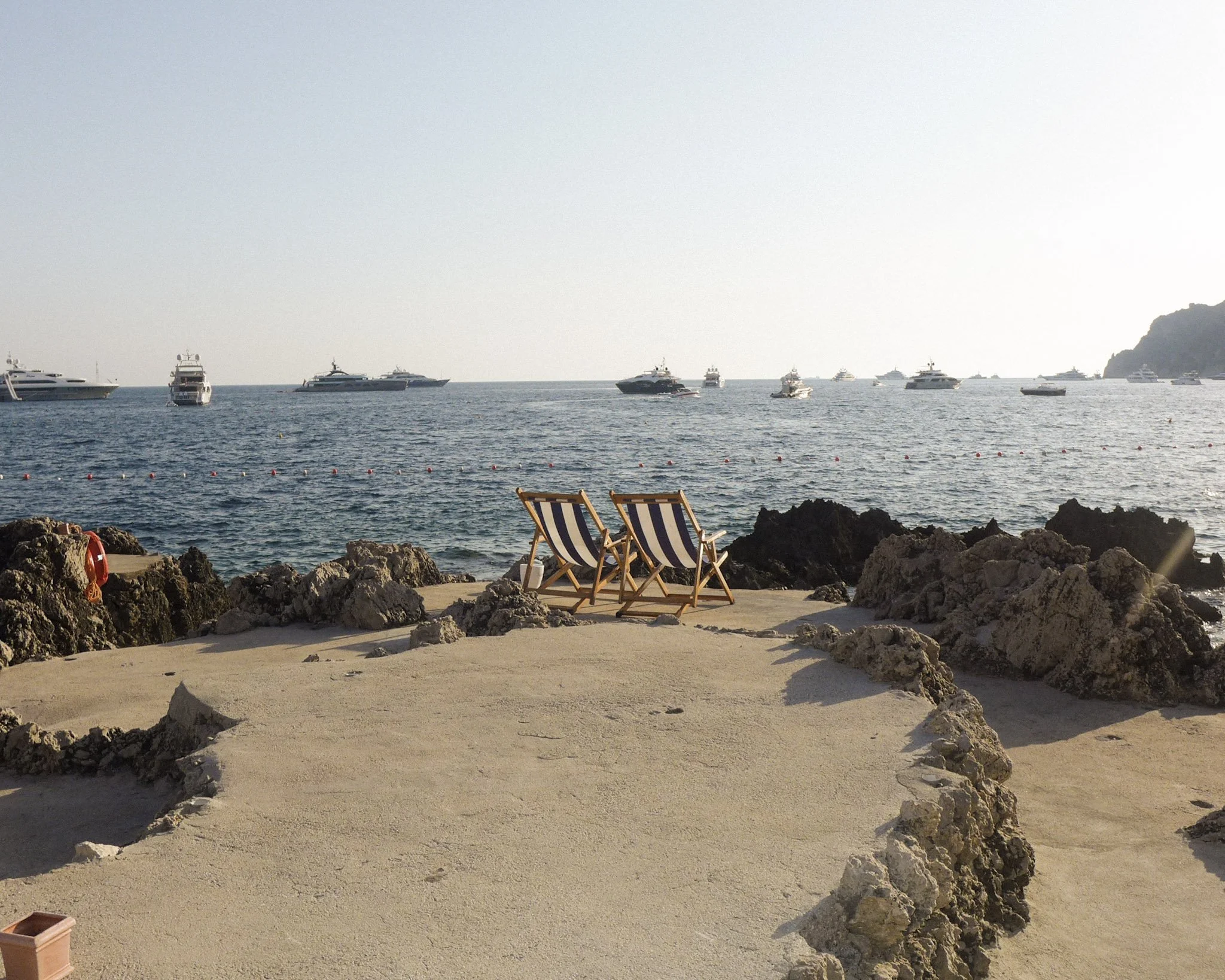 A rocky beach with two striped deck chairs facing the sea, several yachts and boats in the water, a distant cliff on the right, and a clear sky.