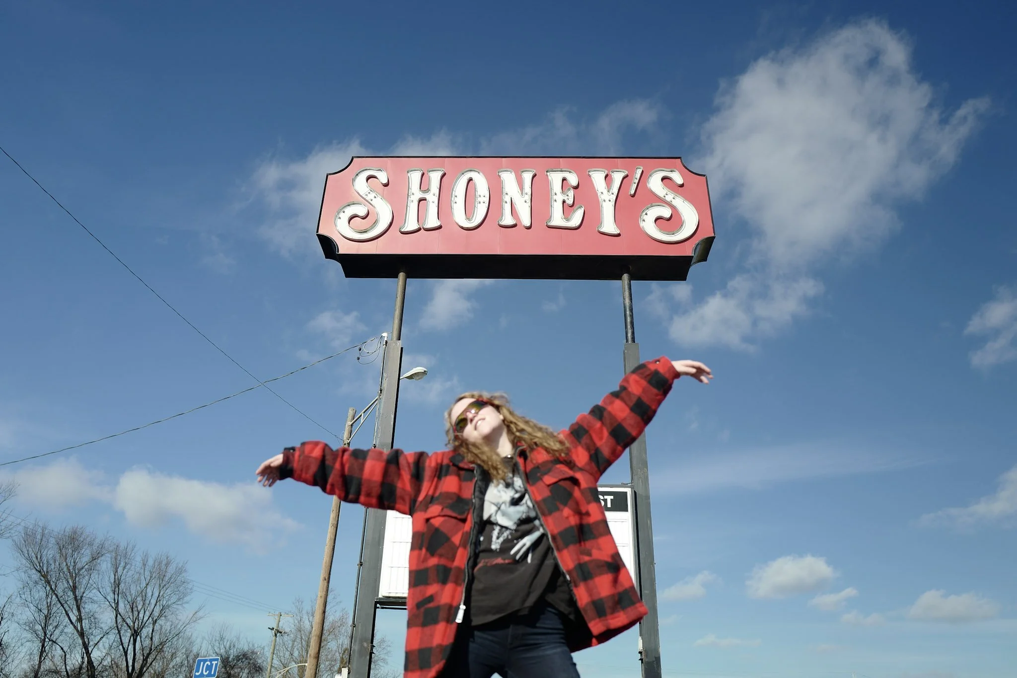 A woman with curly hair wearing sunglasses and a red plaid jacket is standing outdoors with her arms outstretched. Behind her is a pink sign reading 'SHONEY'S' against a blue sky with some clouds.