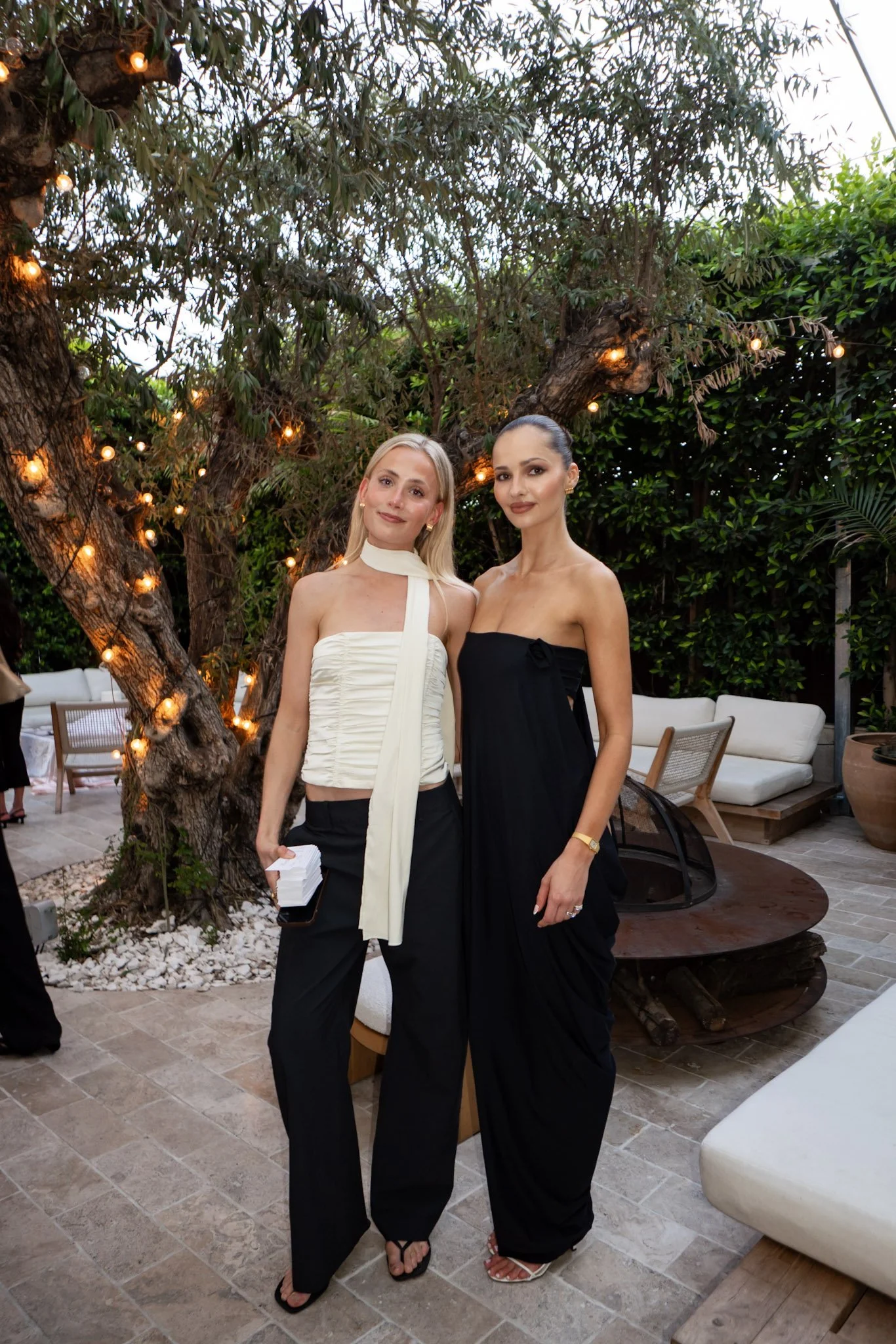 Two women in stylish evening wear stand outdoors under a tree with string lights, with modern patio furniture and greenery in the background.