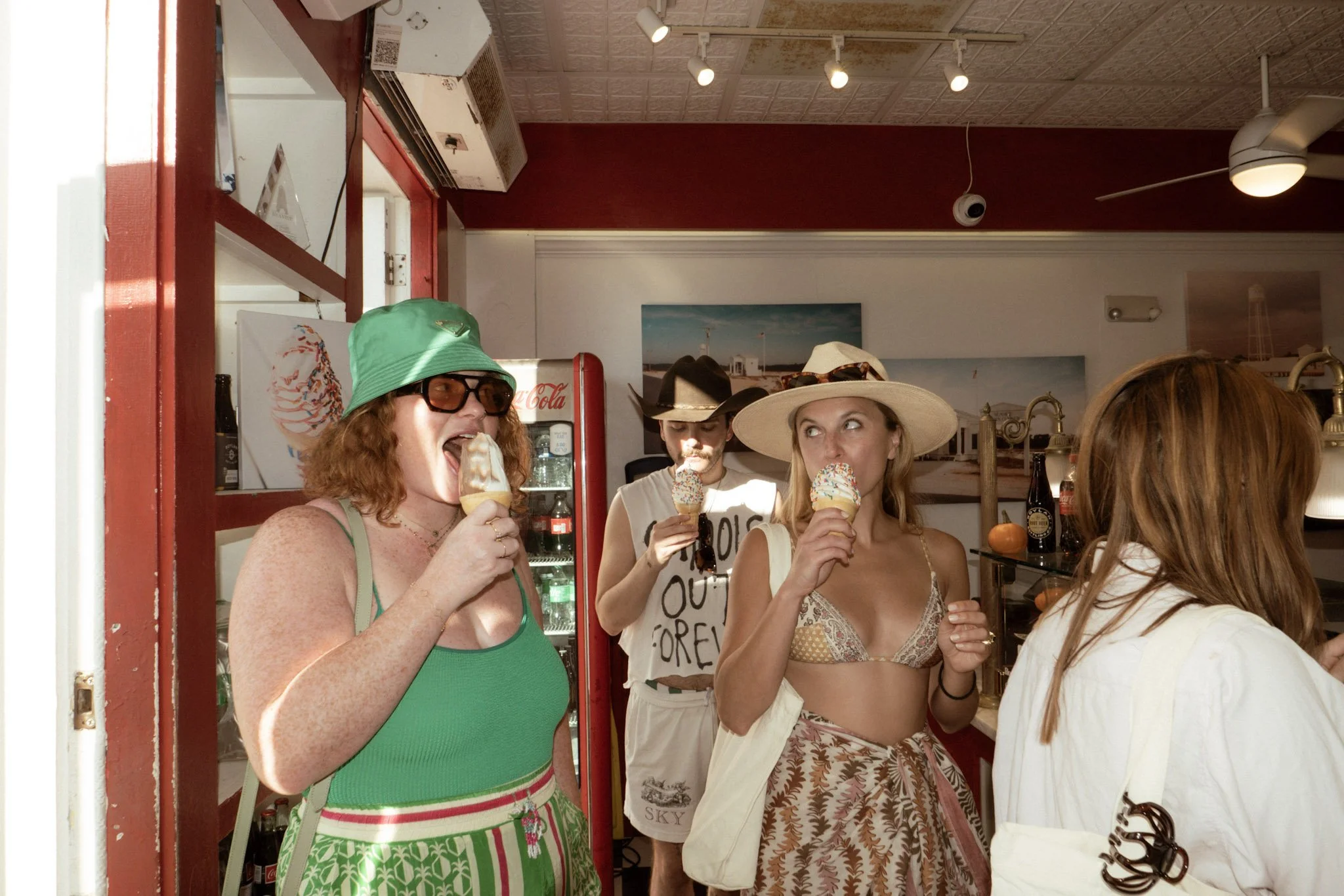 Four women of different ages enjoying ice cream in a casual, tropical-themed restaurant. One woman with curly red hair, wearing sunglasses and a green hat, is licking her ice cream. Another woman with blond hair, wearing a large sun hat, is looking a