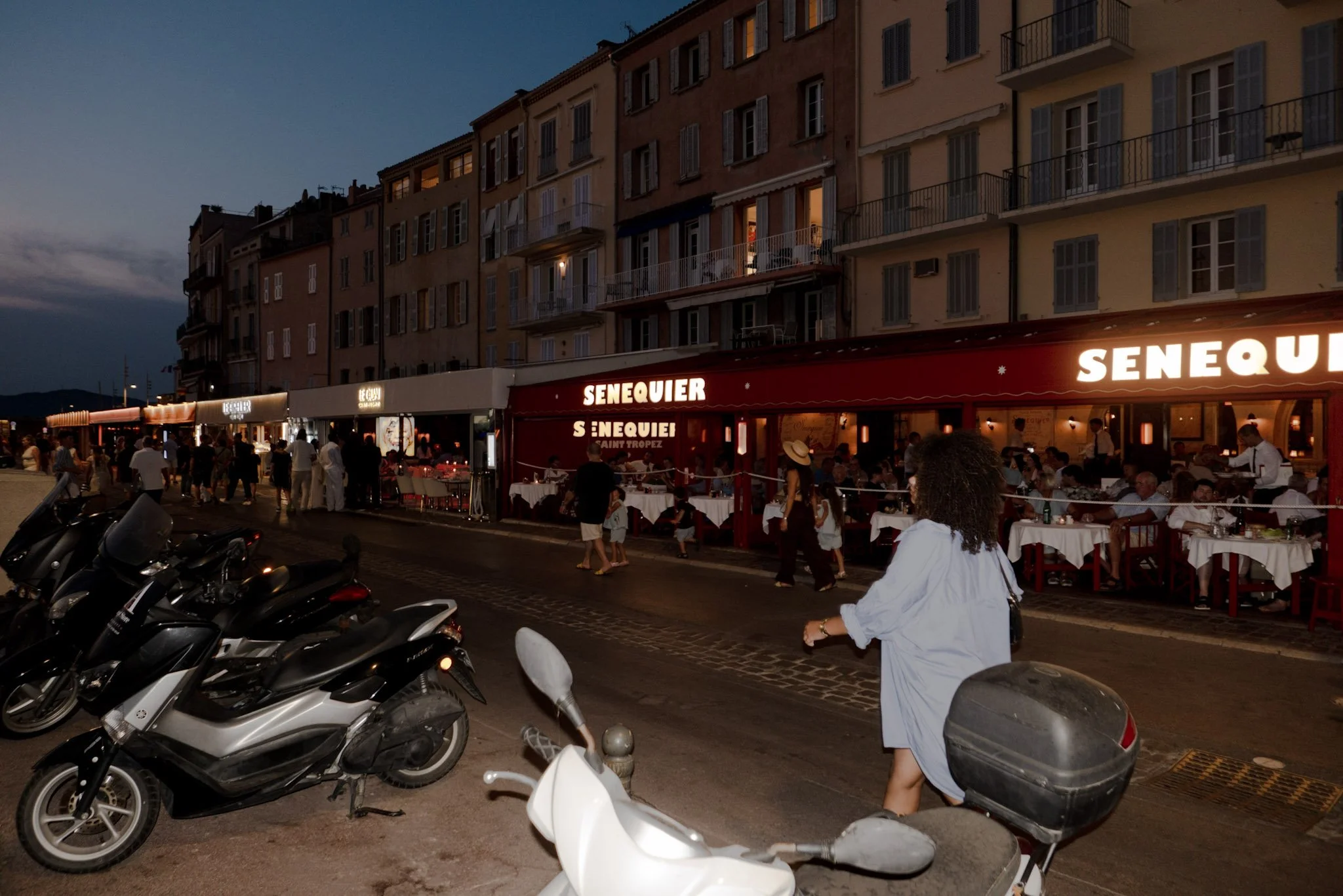 Night scene of a busy street with parked scooters in the foreground, outdoor restaurant with tables and people dining, and a row of tall buildings with illuminated windows and balconies in the background.