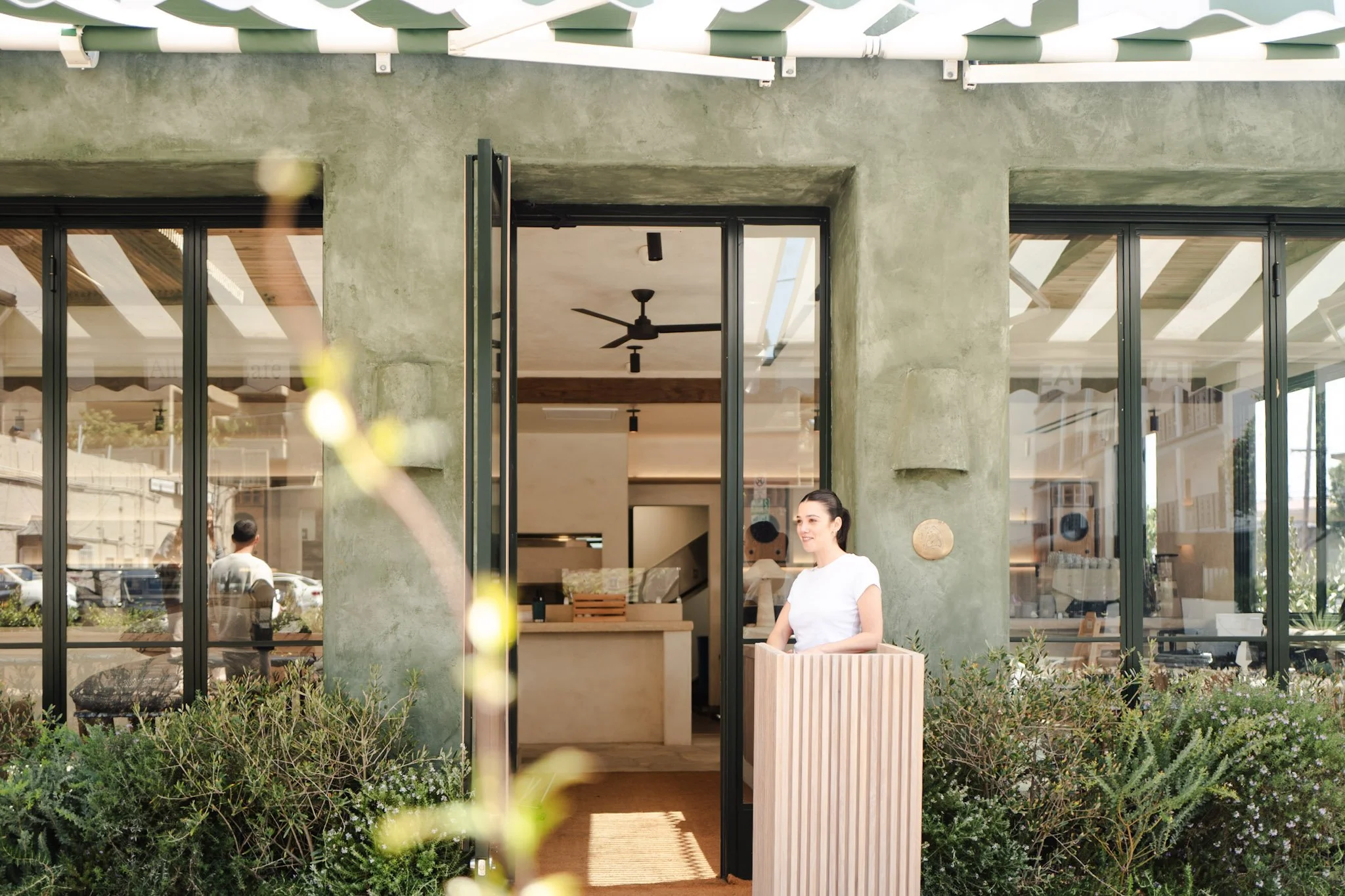 Front view of a modern cafe or restaurant with a concrete exterior and large glass windows. A woman in a white shirt stands outside near a wooden planter box. Inside, ceiling fans are visible through the open doors, and people are dining.