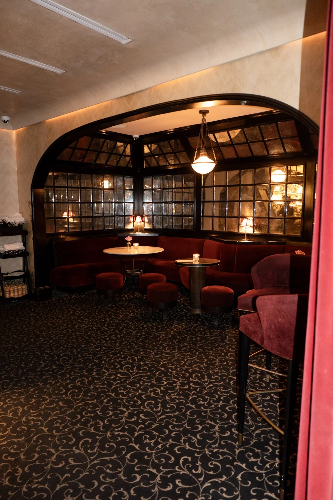 Cozy lounge area with red velvet seating, dark wood tables, small lamps, and a large window with black frames. The room features warm lighting, patterned carpet, and a chandelier hanging from the ceiling.