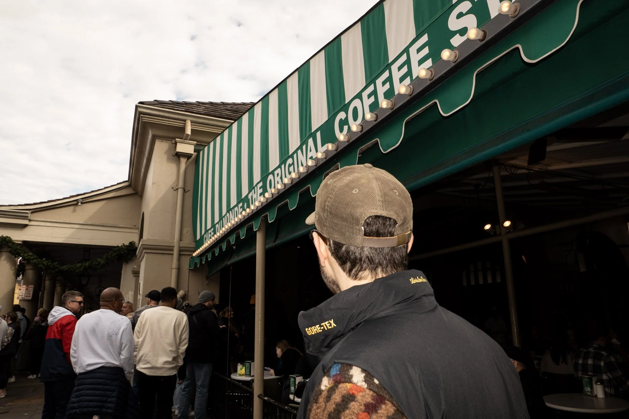 Crowd of people standing outside a building with a green and white striped awning that reads 'The Original' and appears to be a popular food or beverage stand.