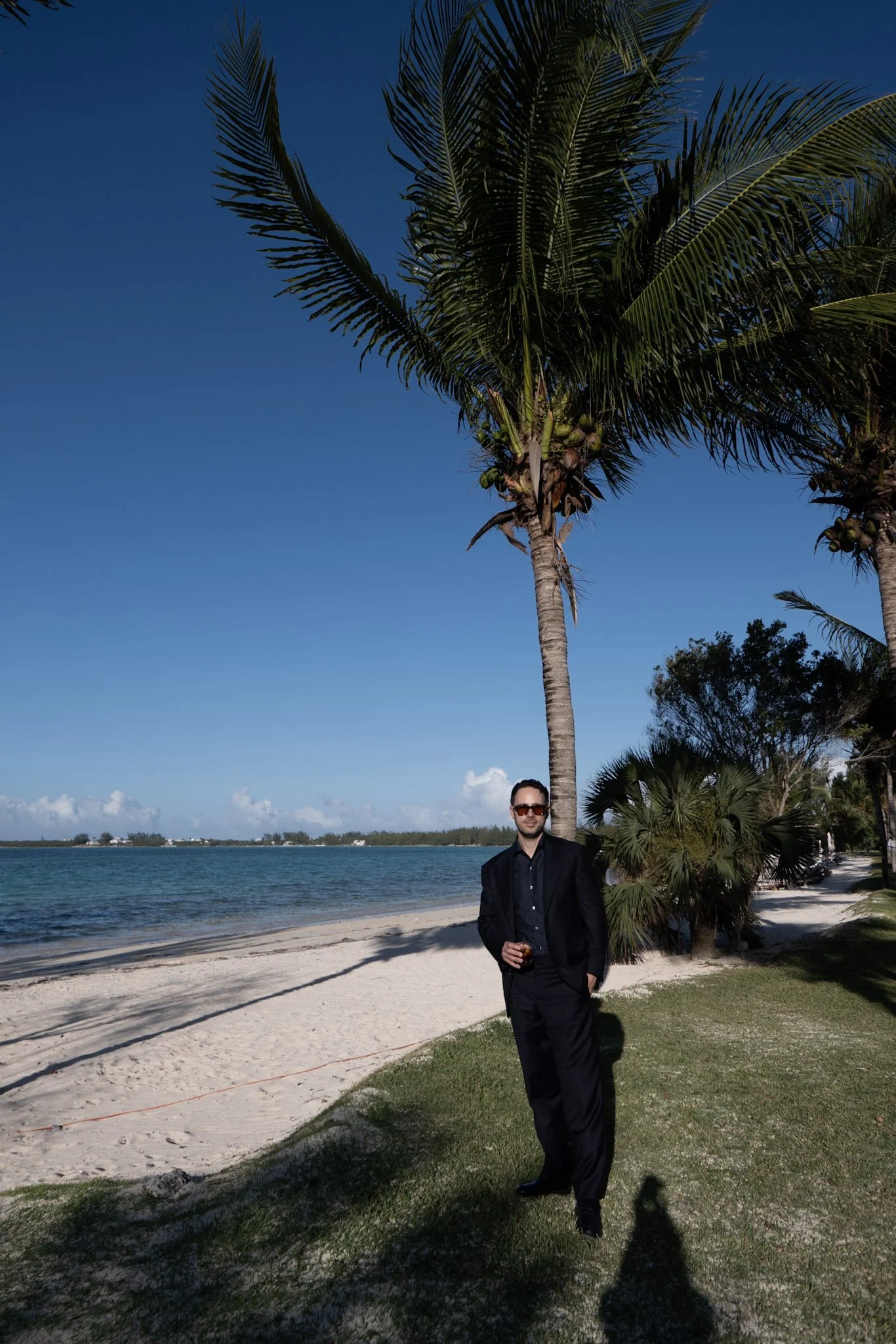 A man in a black suit and sunglasses standing on a sandy beach with palm trees, a grassy area, and a body of water with a clear blue sky in the background.