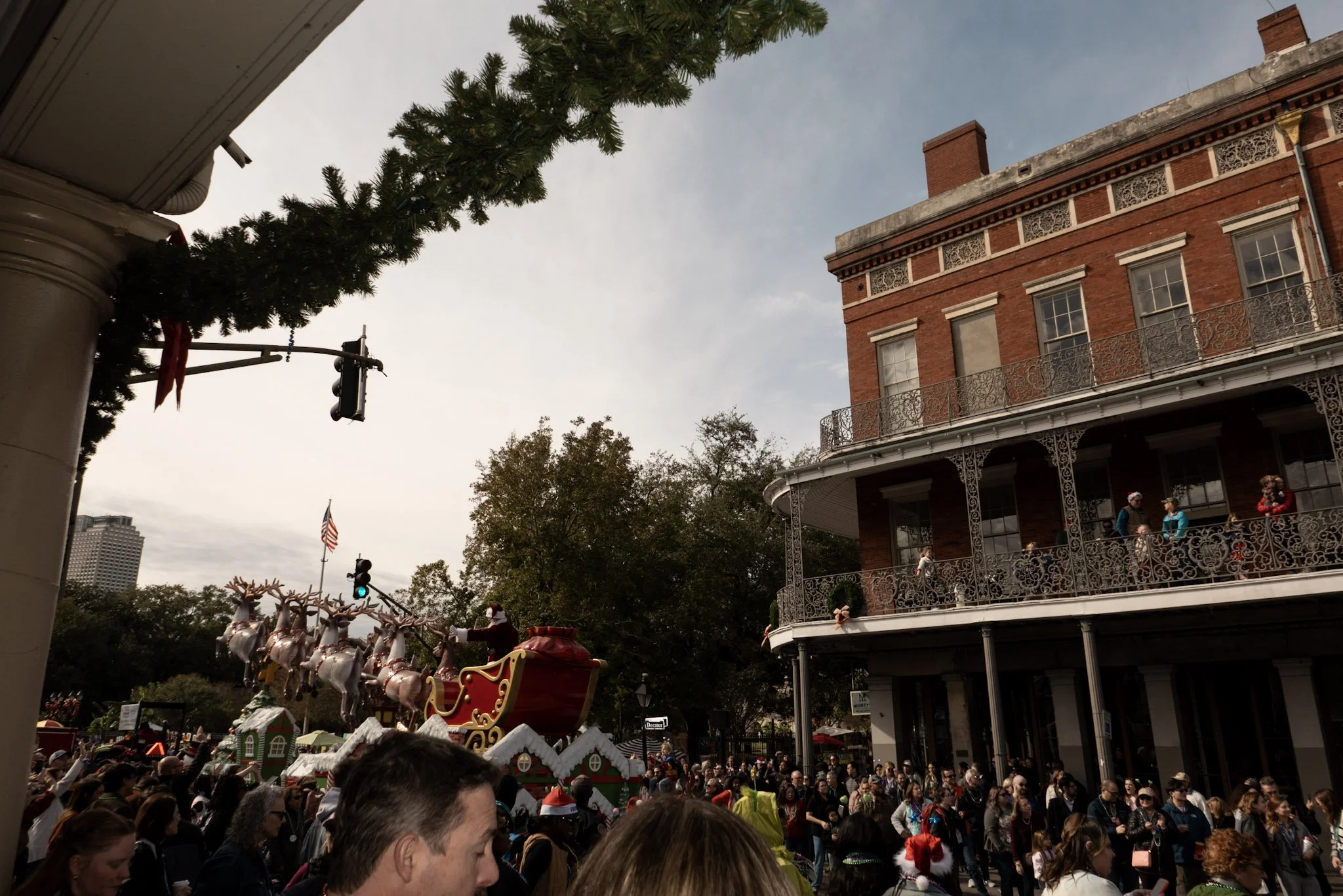 A crowded street scene with people lining up for a festive event, featuring a large building with wrought iron balconies, Christmas decorations, and a festive parade with Santa's sleigh and reindeer.