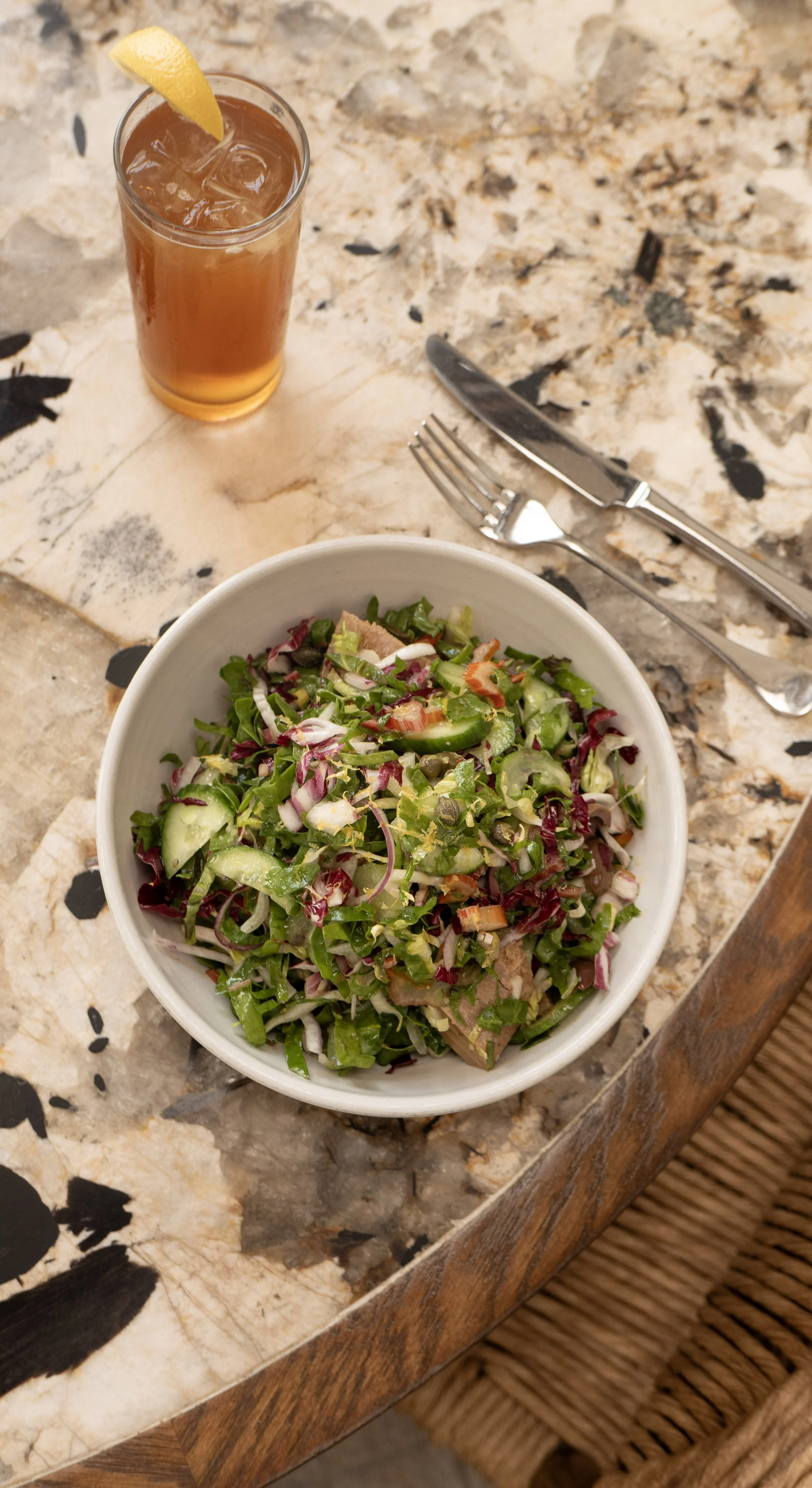 A bowl of mixed salad with cucumbers, tomatoes, onions, lettuce, and capers, on a granite table with silverware, and a glass of iced tea with lemon.