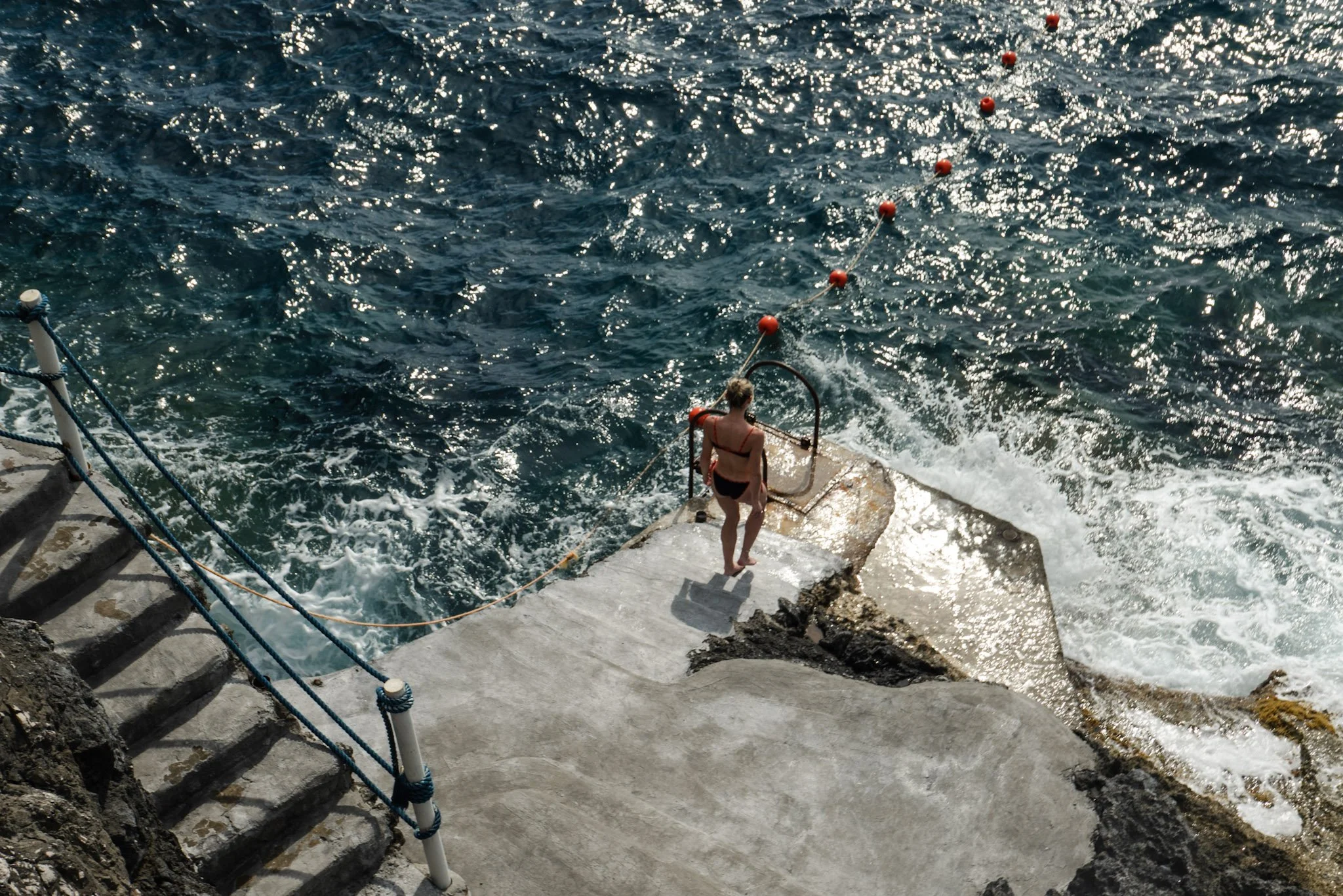 A woman in a bikini standing on a concrete pier at the water's edge, holding onto a metal railing with orange buoys floating in the water nearby.