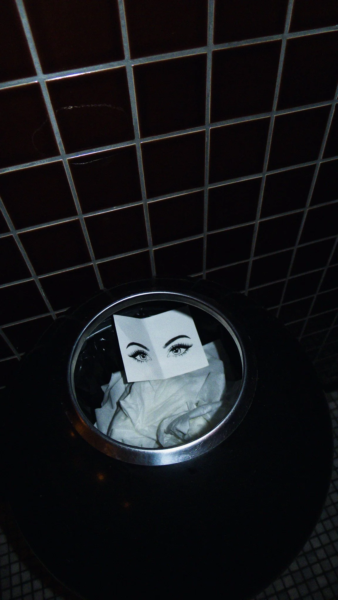 A black trash can with a piece of paper featuring a drawing of eyes on top, inside the trash can. The background consists of black tiled wall.