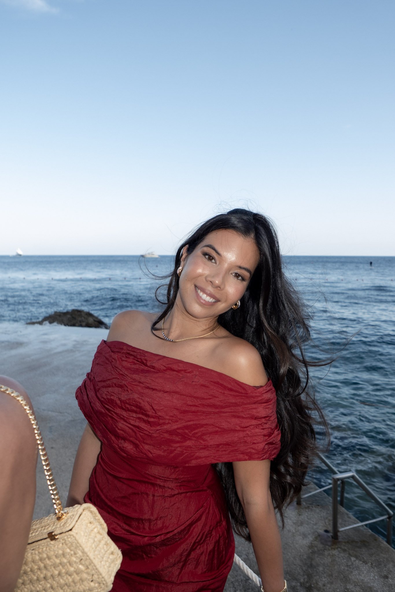 A woman with long dark hair smiling at the camera at a beach, wearing an off-the-shoulder red dress, with sea and boats in the background.