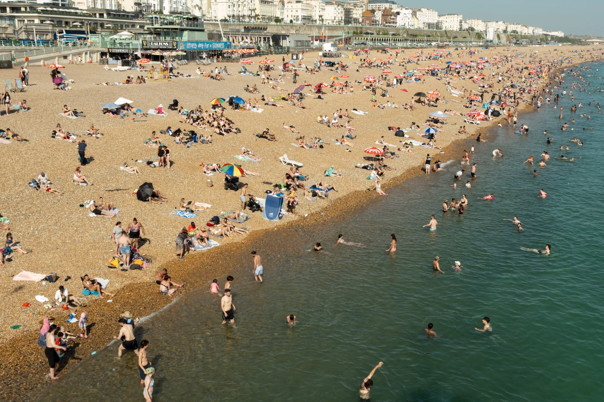 Crowded beach with people sunbathing, swimming, and walking along the shoreline, umbrellas and beach chairs scattered across sandy area, and white buildings in the background.