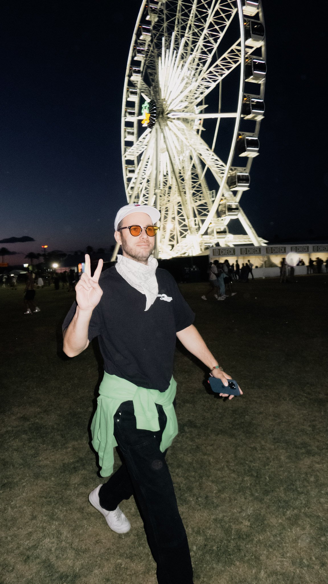 A man wearing sunglasses and a white cap making a peace sign while walking in front of a large Ferris wheel at night.