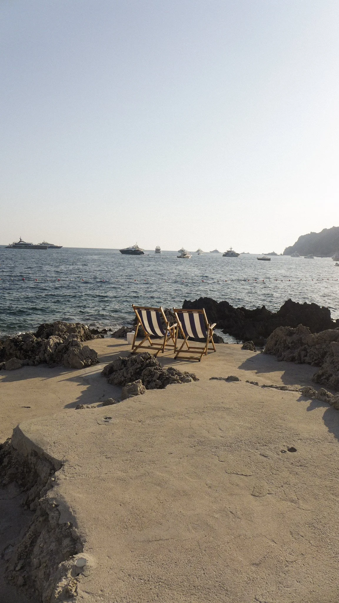 Two striped lounge chairs on a sandy beach next to rocks, facing the ocean with several boats in the water and a distant landmass on the horizon.