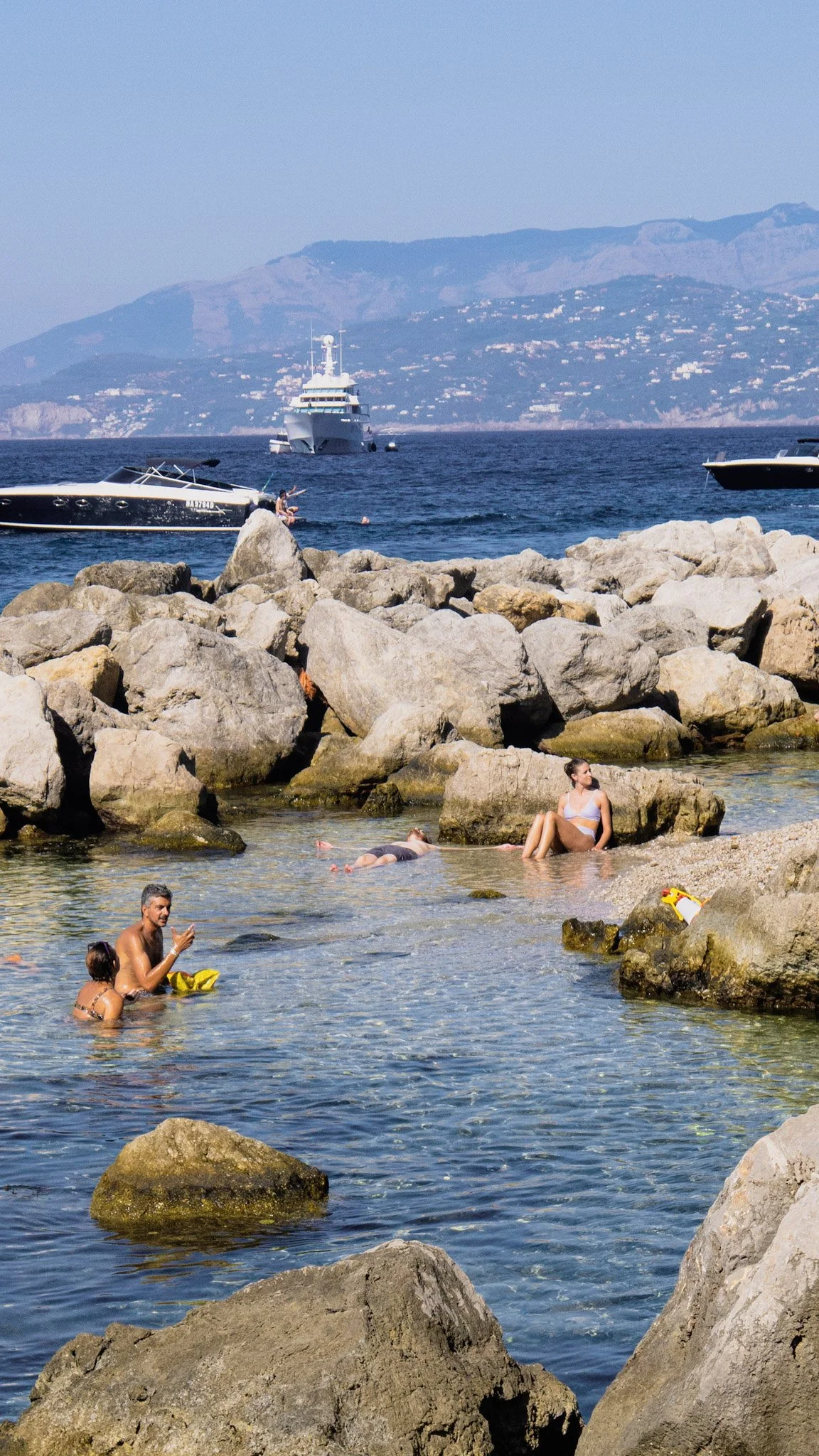 People relaxing and swimming near rocks at a beach, with boats and a mountain in the background.