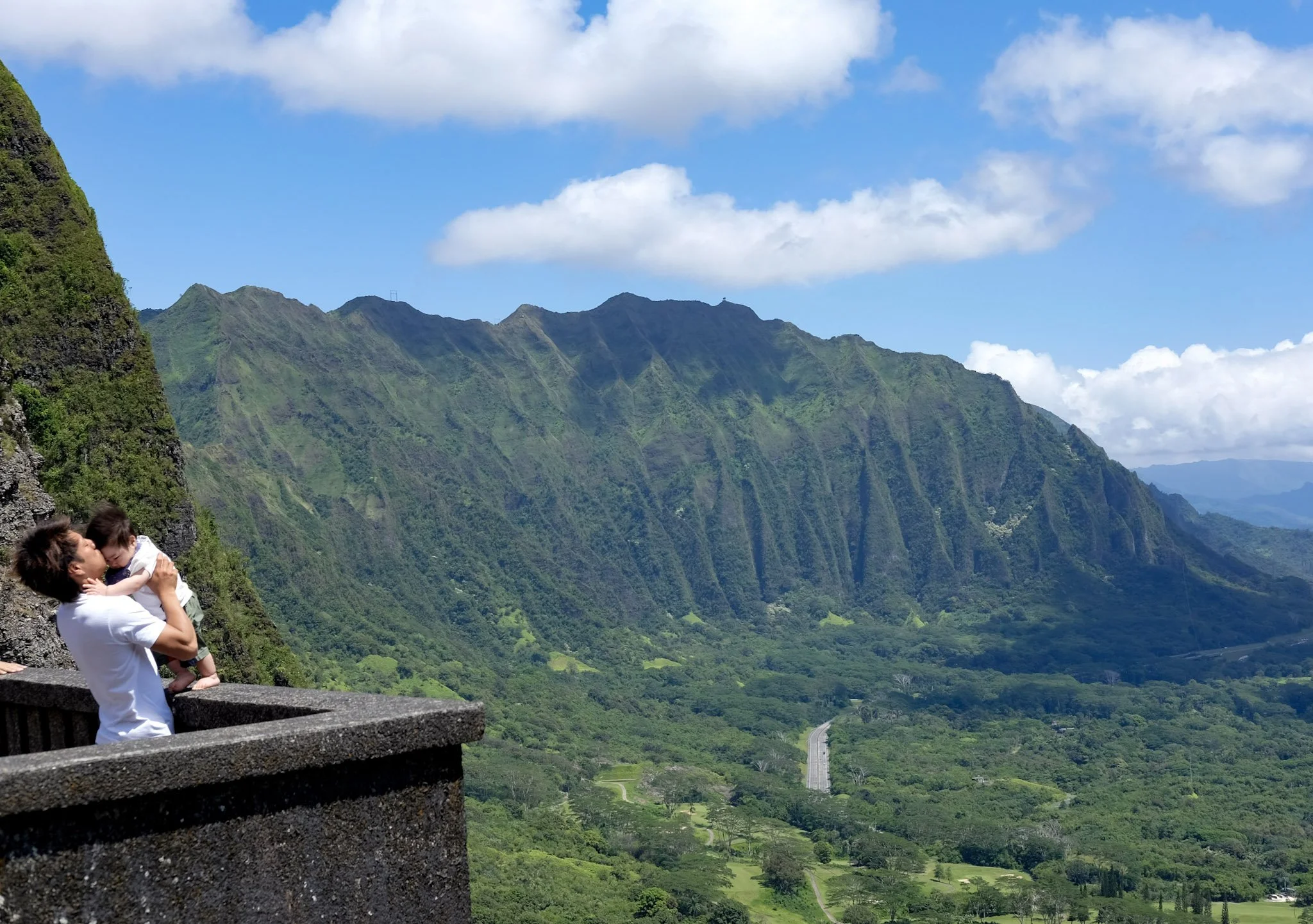 A person holding a child on a stone viewing platform overlooking lush green mountains and a valley with a road, under a partly cloudy sky.