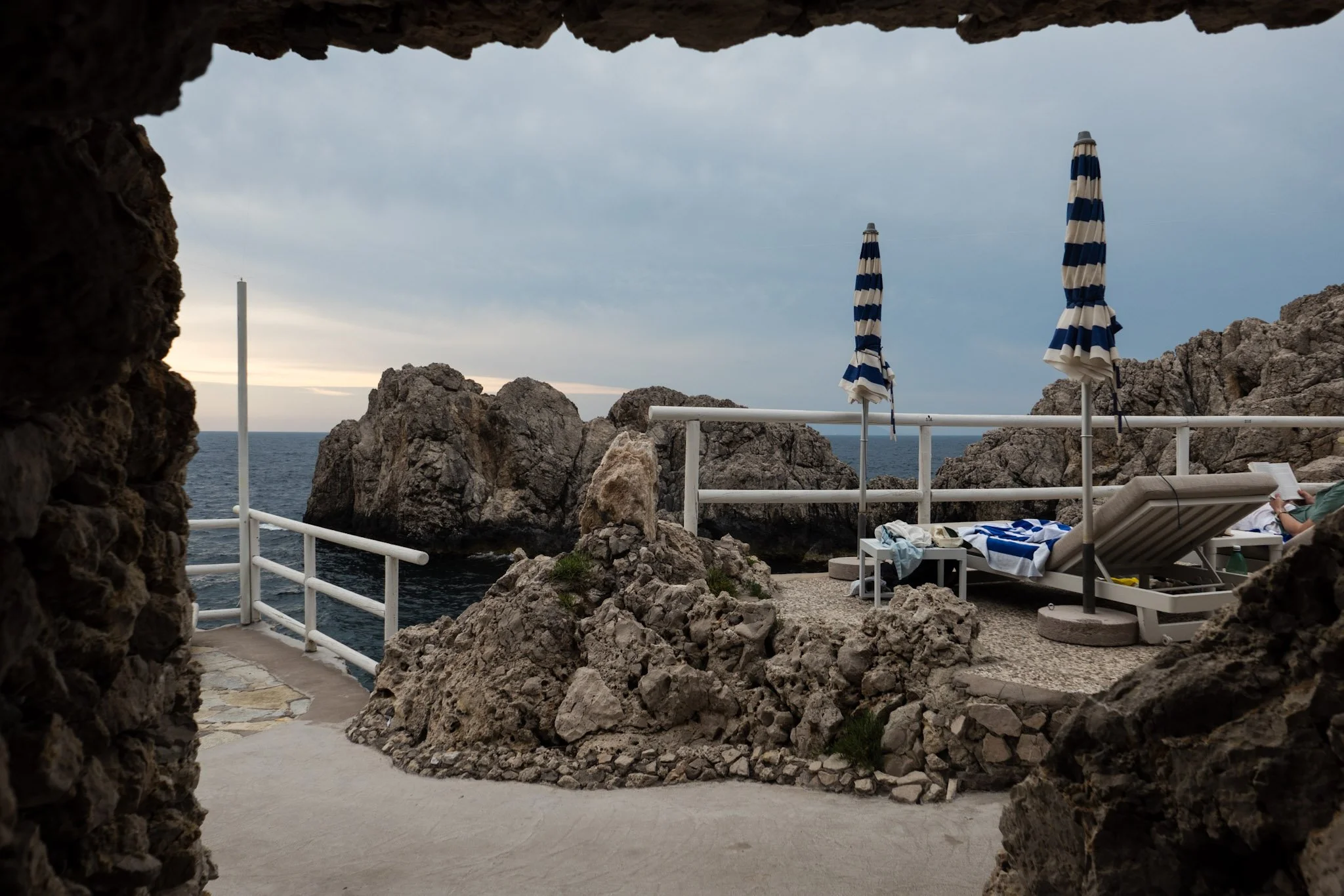 View of a rocky seaside area with lounge chairs and striped umbrellas, seen through a natural stone archway, overlooking the ocean under a cloudy sky.