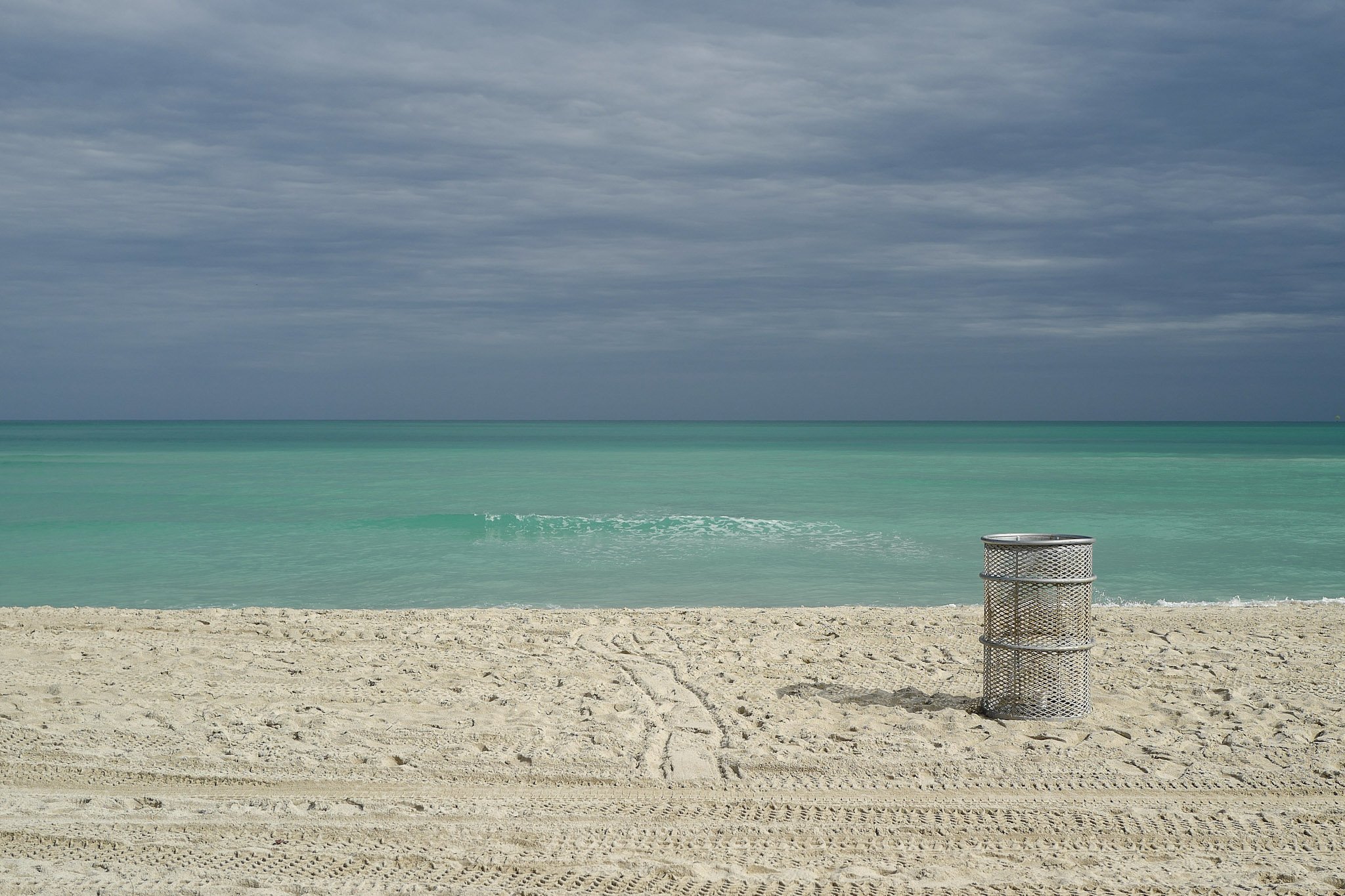Empty beach with a trash can and cloudy sky over turquoise ocean.