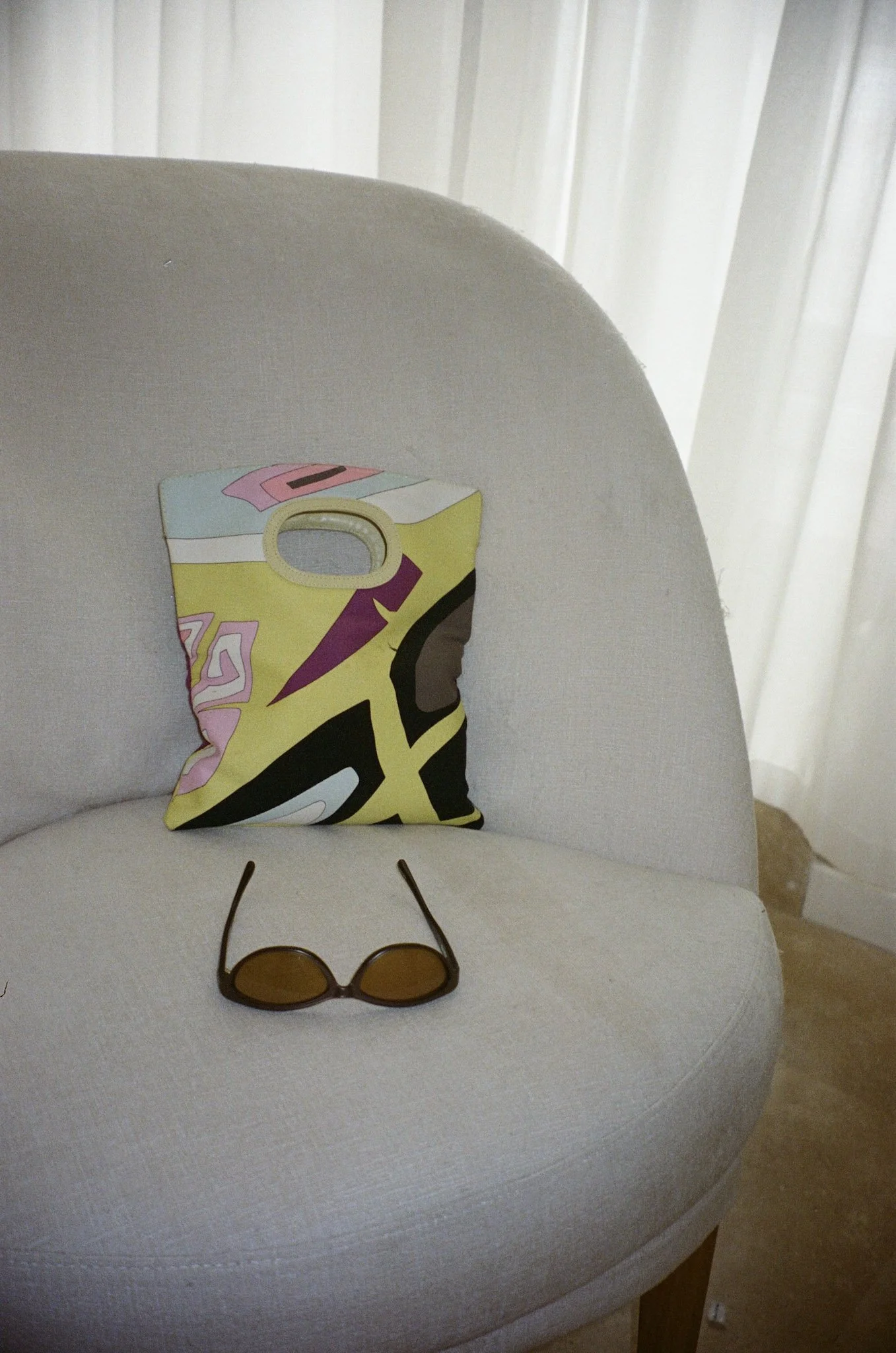 A white upholstered chair with a colorful foldable paper mask, a pair of sunglasses with dark brown lenses, and white vertical blinds in the background.
