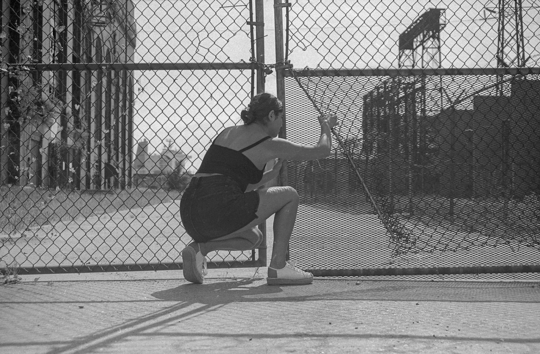 A woman squatting on a chair behind a chain-link fence, holding the fence with one hand and looking to the side in an outdoor urban setting. Black and white photo.