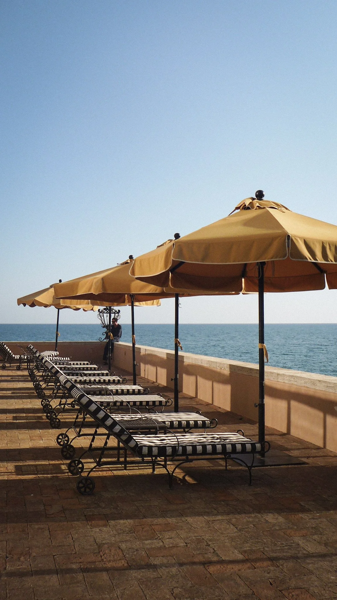 Beachside outdoor lounge area with striped lounge chairs under large yellow umbrellas, overlooking the ocean with a clear blue sky.