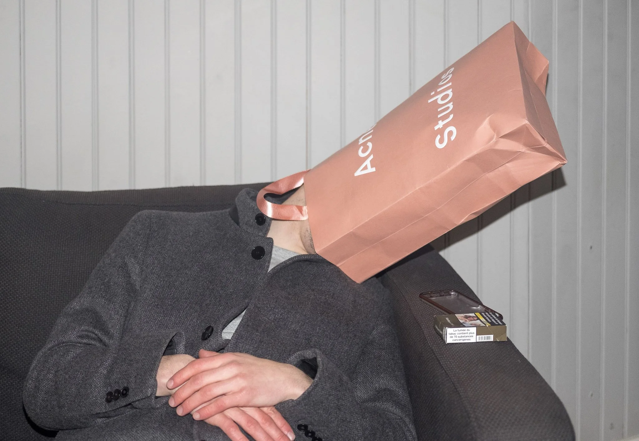 Person dressed in a gray coat and gray shirt, holding a pink shopping bag with white text, standing in front of a white panel wall, with a pack of cigarettes placed on a brown surface next to them.