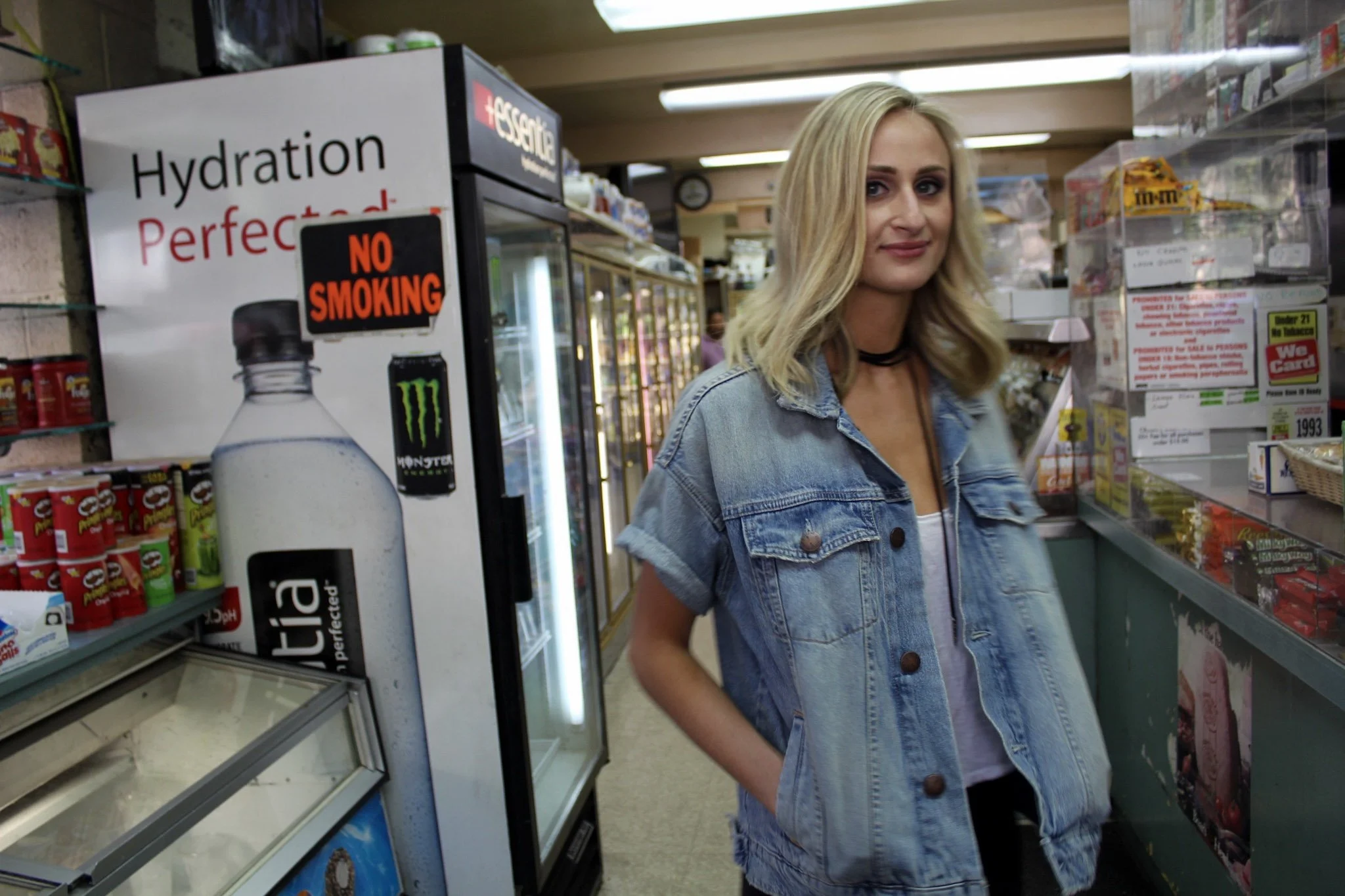 A woman with blonde hair wearing a denim jacket and black choker shopping in a store aisle with shelves stocked with snacks and beverages, including a soda fridge in the background.