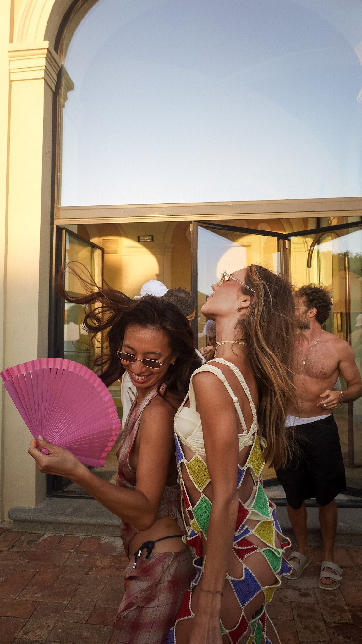A group of young people dancing and enjoying themselves outside a building, with women wearing colorful outfits and holding a pink fan.