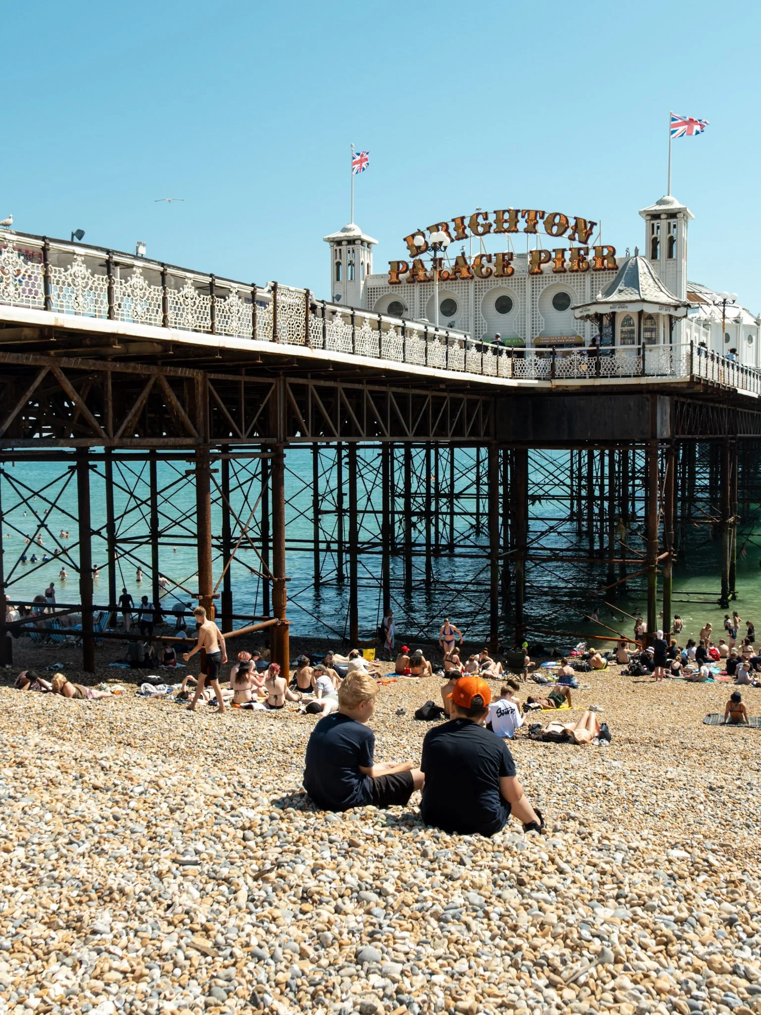 People relaxing and sunbathing on a rocky beach under the Brighton Palace Pier in Brighton, England, with the pier's white structure and flags visible in the background.