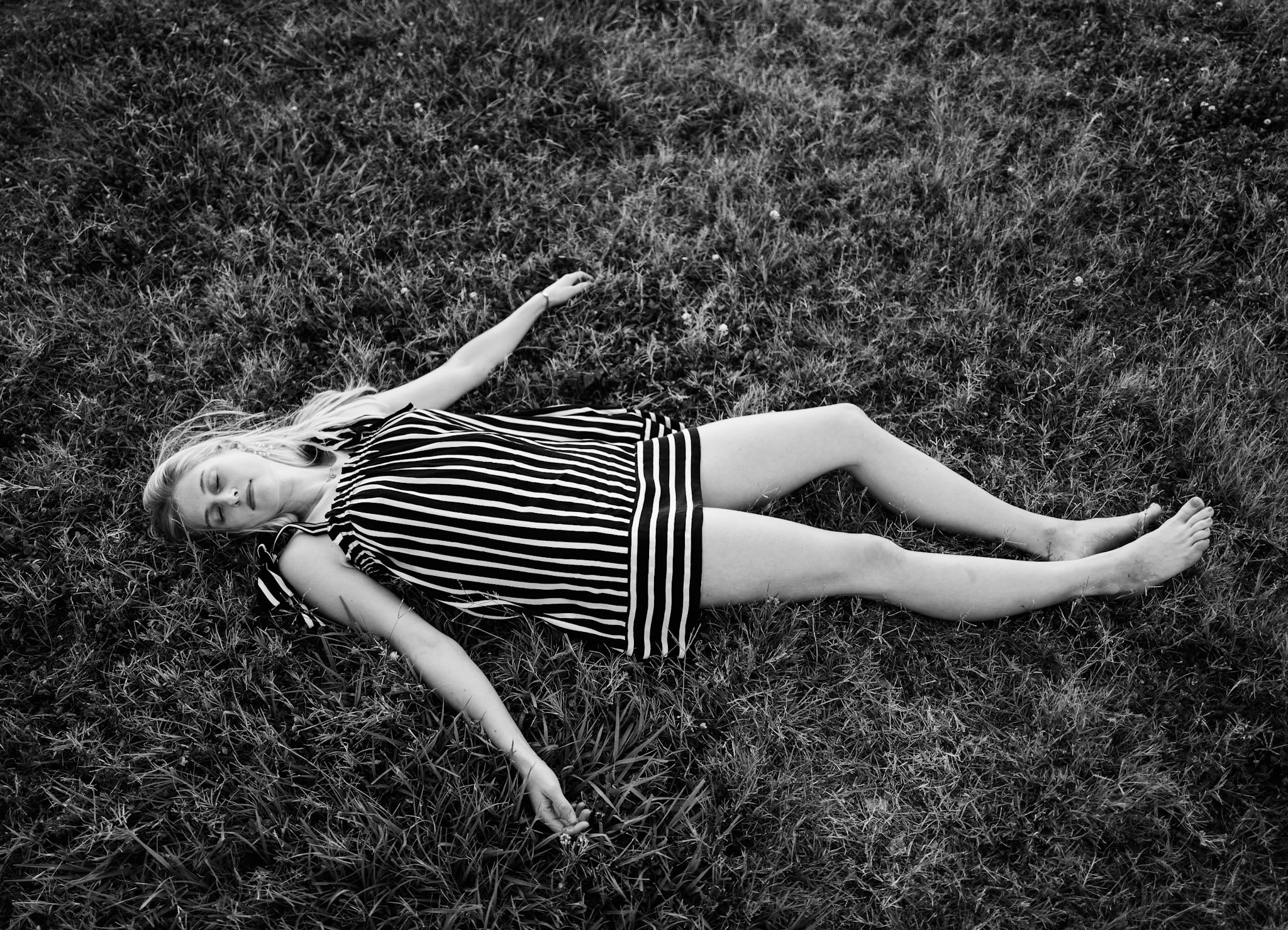 A young girl lying on her back on grass with her eyes closed, wearing a striped shirt and shorts, in a black and white photograph.