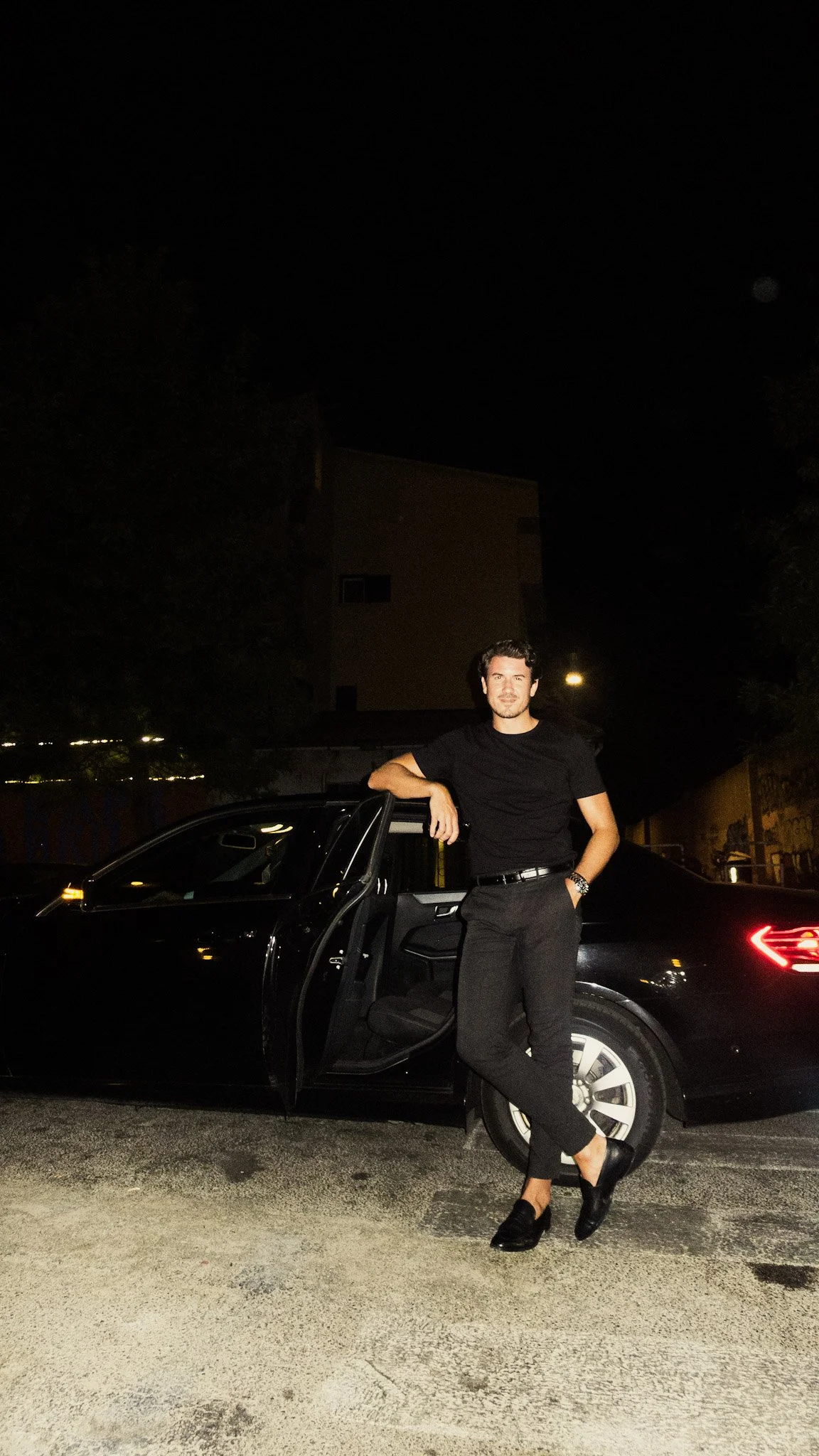 A man leaning against a black car with the driver's door open, standing outside at night, dressed in black pants and shoes, with a black t-shirt, in front of a building with trees around, illuminated by nearby streetlights.