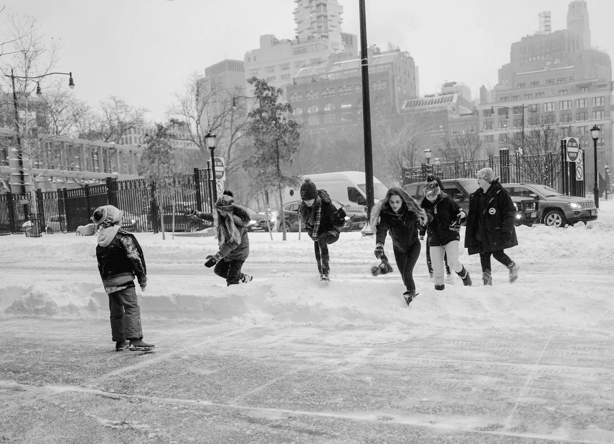 Children and a woman walking in snow-covered park with tall city buildings in background, some trees, street lamps, and cars