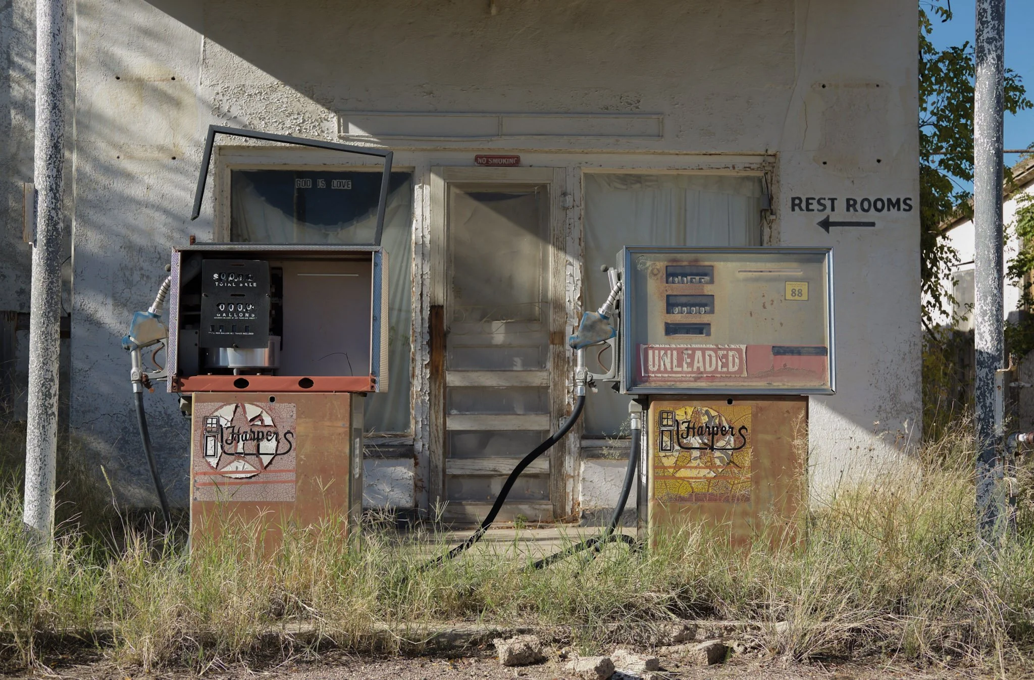 An old, weathered building with a white, peeling exterior, two gas pumps in front, one labeled "Unleaded," with hoses hanging from them. Sign indicating "Rest Rooms" pointing left is on the wall. The building has a small window and a door with a curt