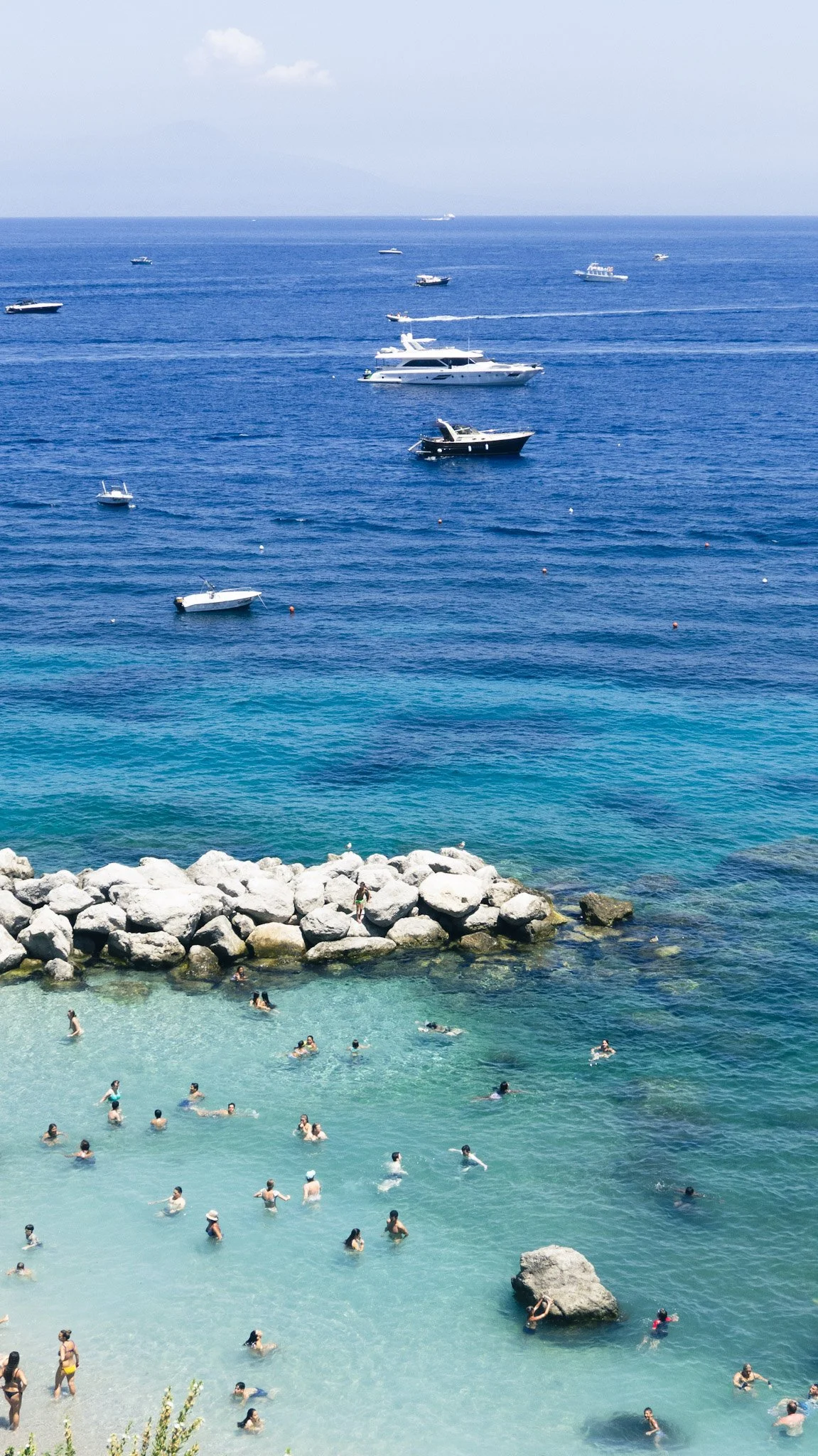 People swimming in a turquoise beach with large rocks, with boats floating on the blue ocean and a mountainous island in the distance.