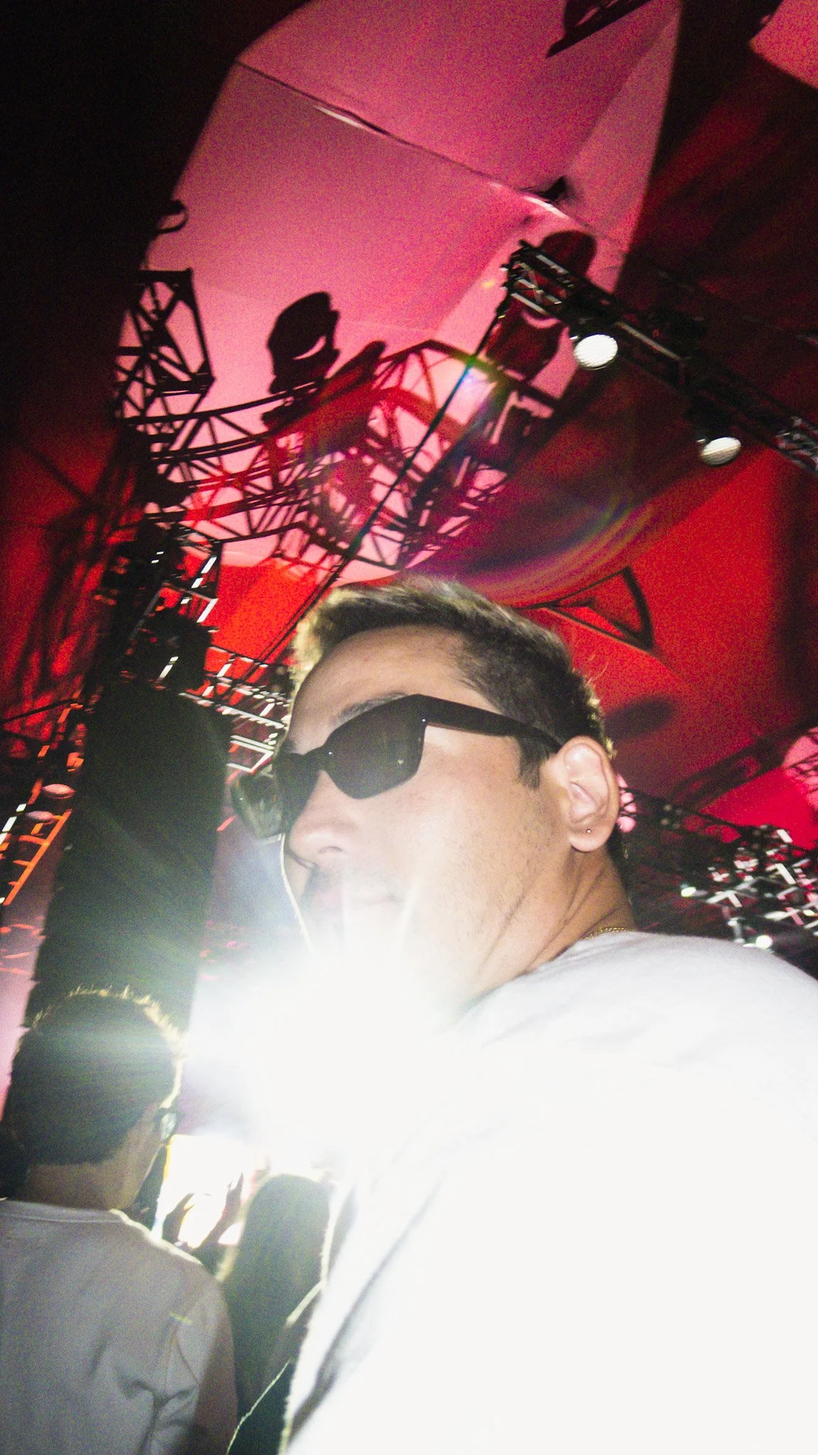 A person wearing sunglasses and a white shirt taking a selfie at a concert with stage lights and scaffolding in the background.
