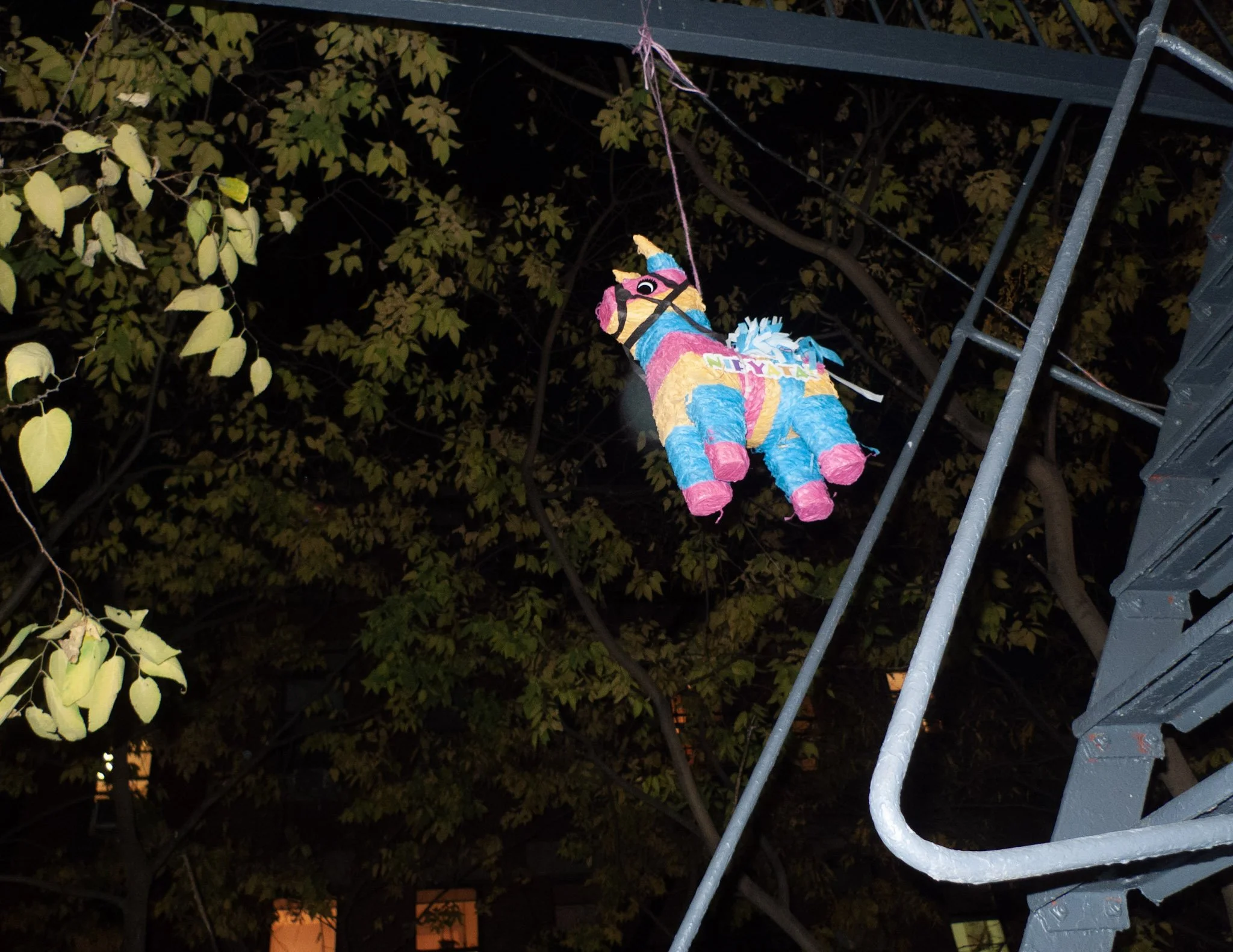 Colorful piñata unicorn hanging from a pink string on a balcony at night with trees in the background.