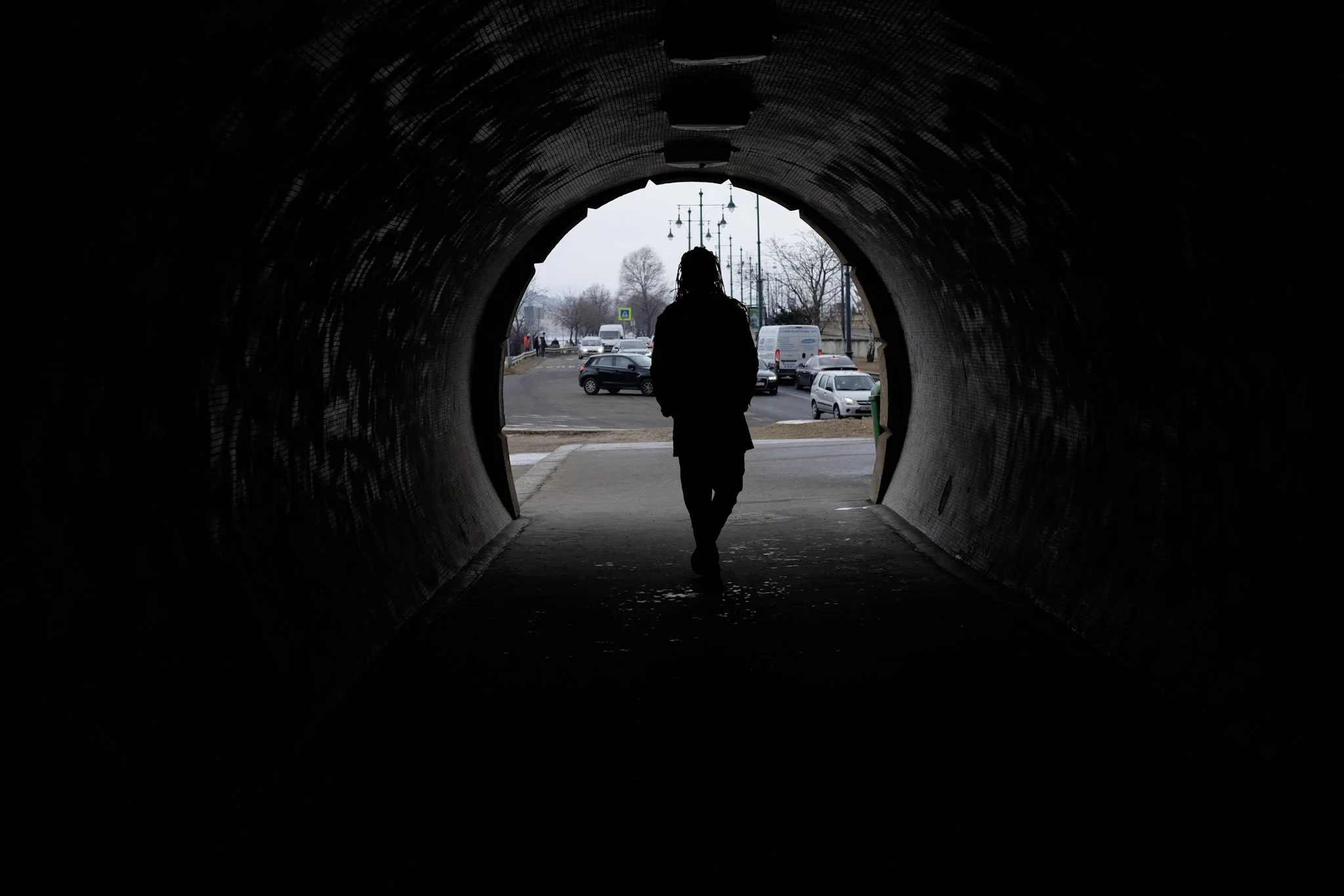A person with dreadlocks walking through a dark tunnel towards a city street with cars and streetlights.