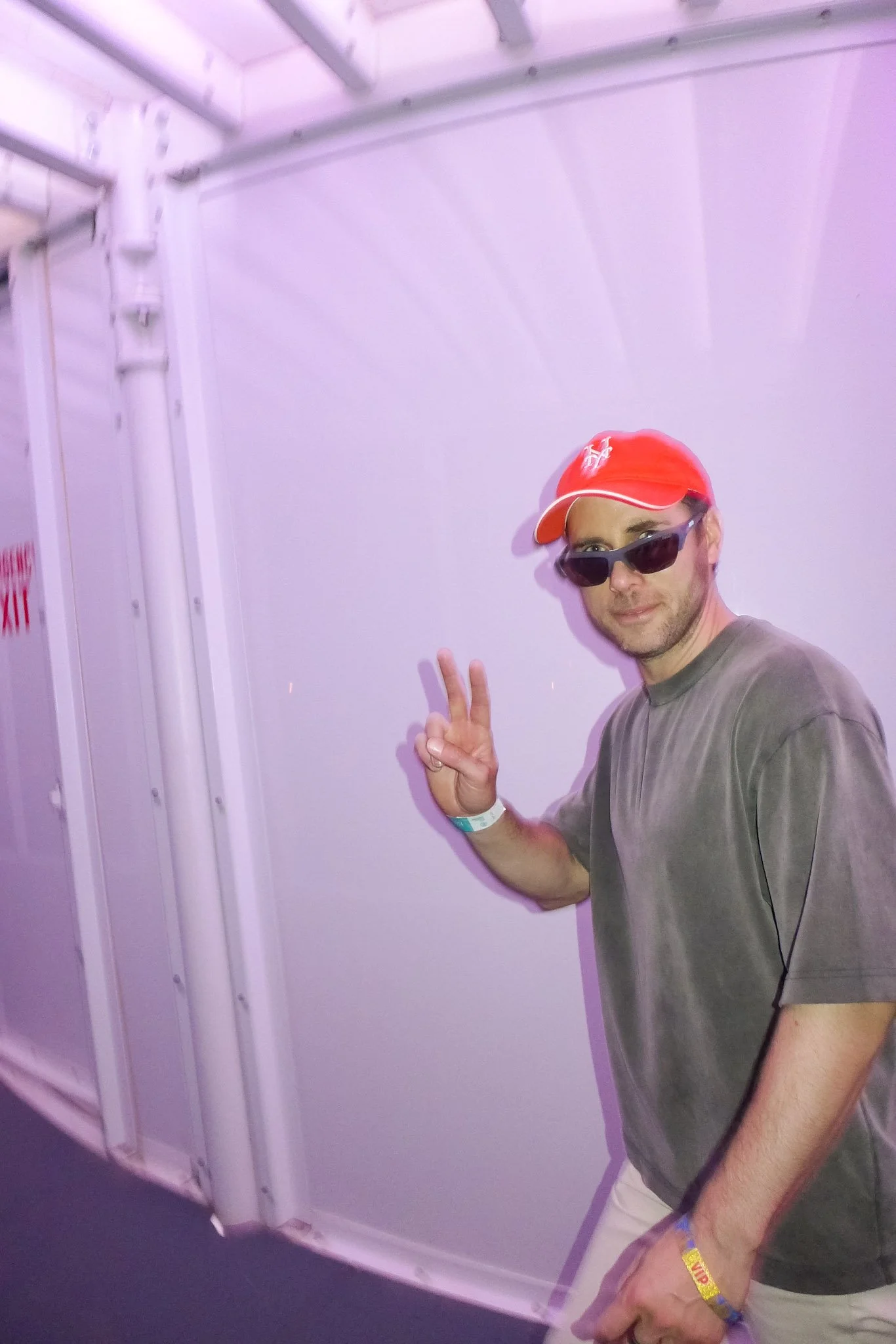 A man wearing a red baseball cap, sunglasses, and a gray t-shirt, holding up a peace sign with his right hand, standing next to a white wall with industrial pipes and a fire escape.