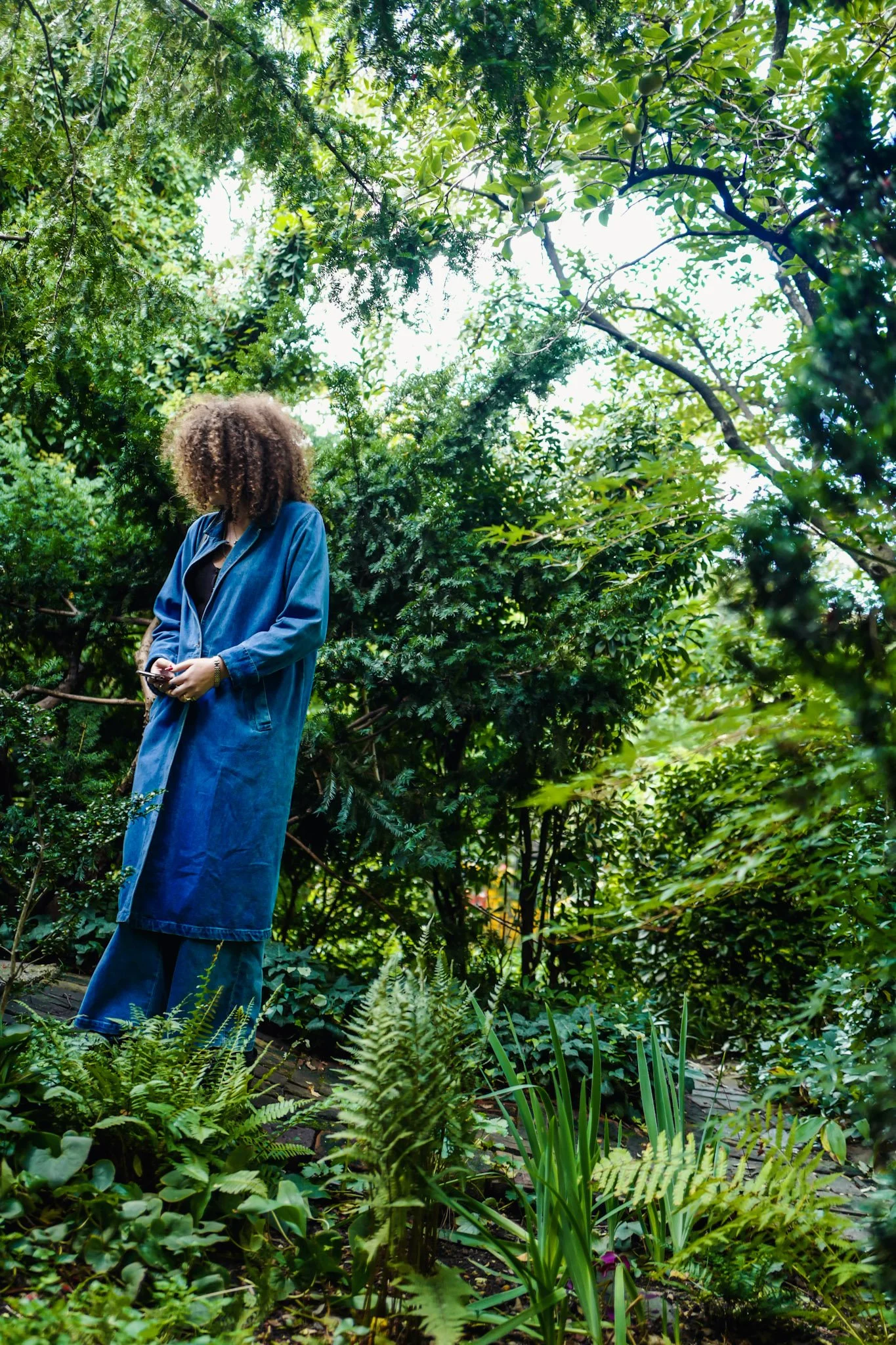 Person with curly hair wearing a blue denim jacket and matching pants, standing in lush green forest, looking at their phone.