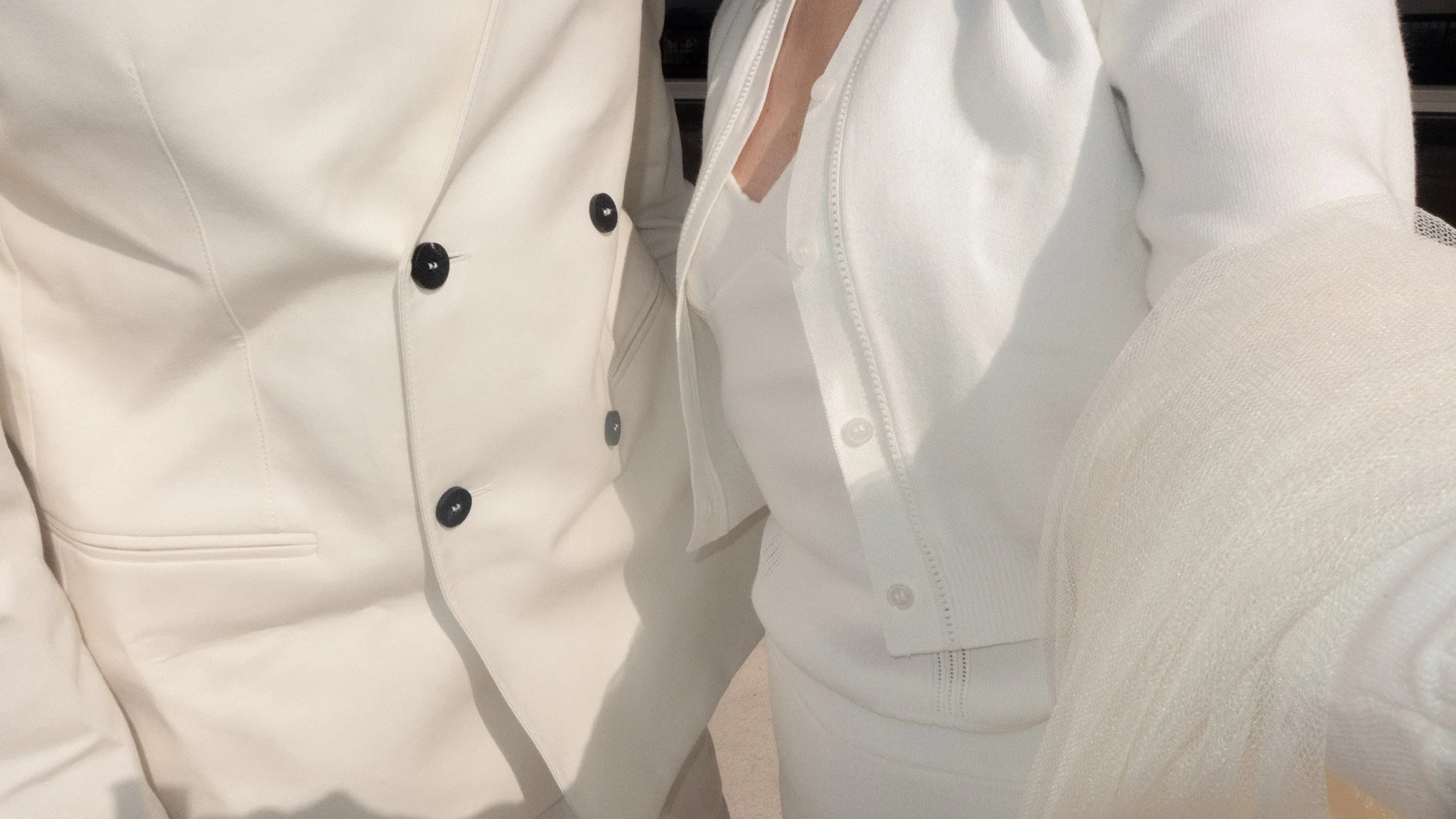 Close-up of a person wearing a white blazer with black buttons and a white top underneath, sitting on a beige couch.