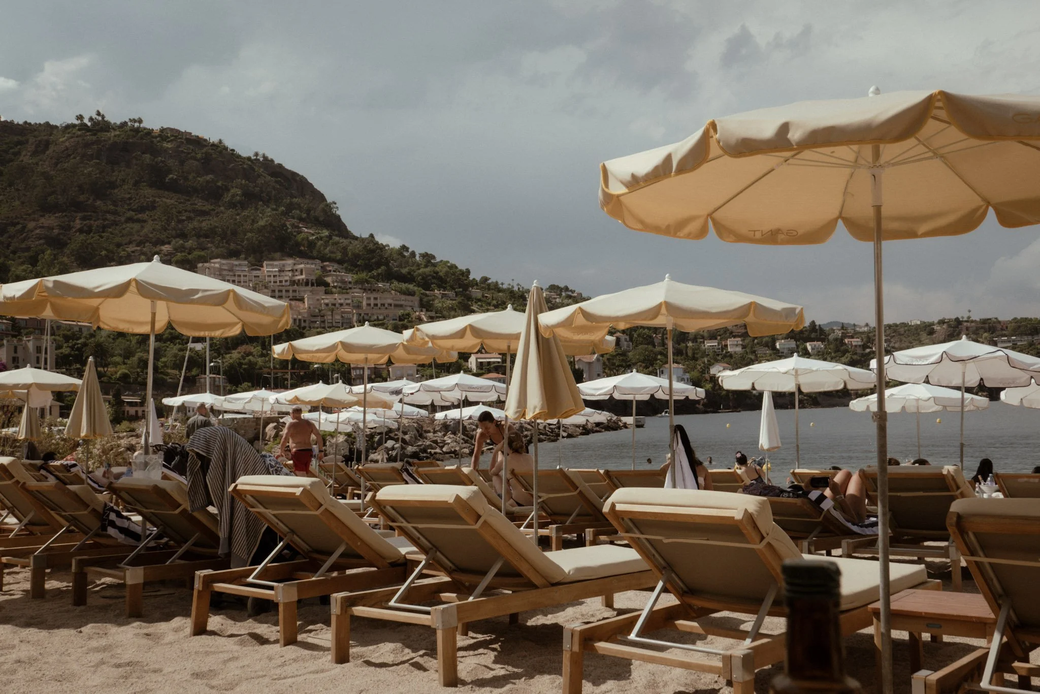 Beach scene with numerous beige umbrellas and lounge chairs along the sandy shore, with people relaxing and walking near the water. Hills with houses and cloudy sky in the background.