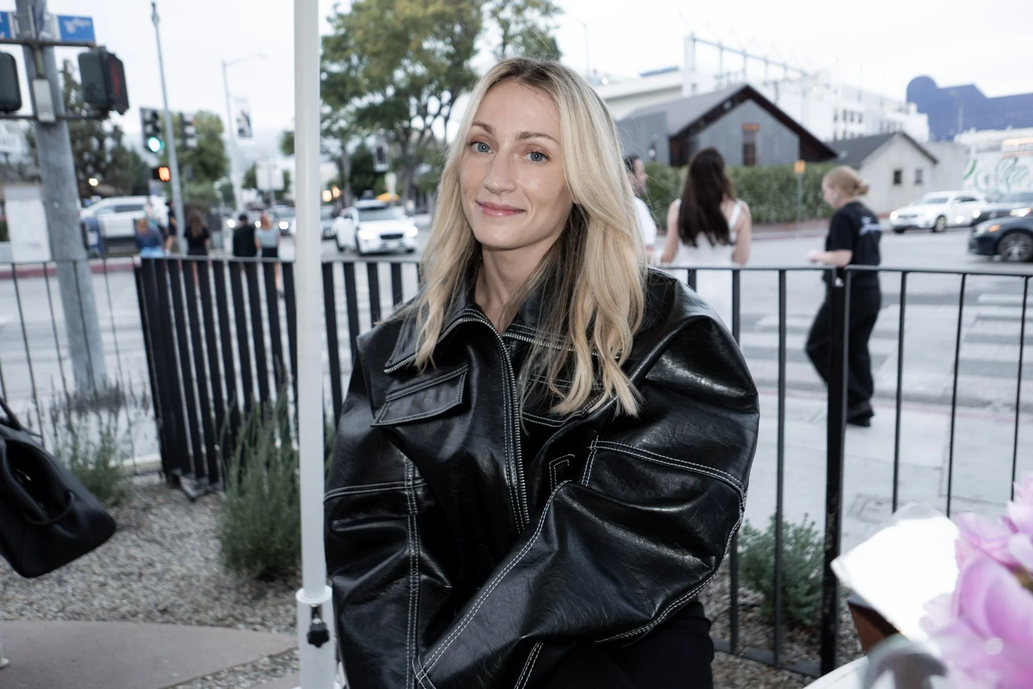 A woman with blonde hair, wearing a black leather jacket, sitting outdoors near a busy intersection with cars and pedestrians in the background.