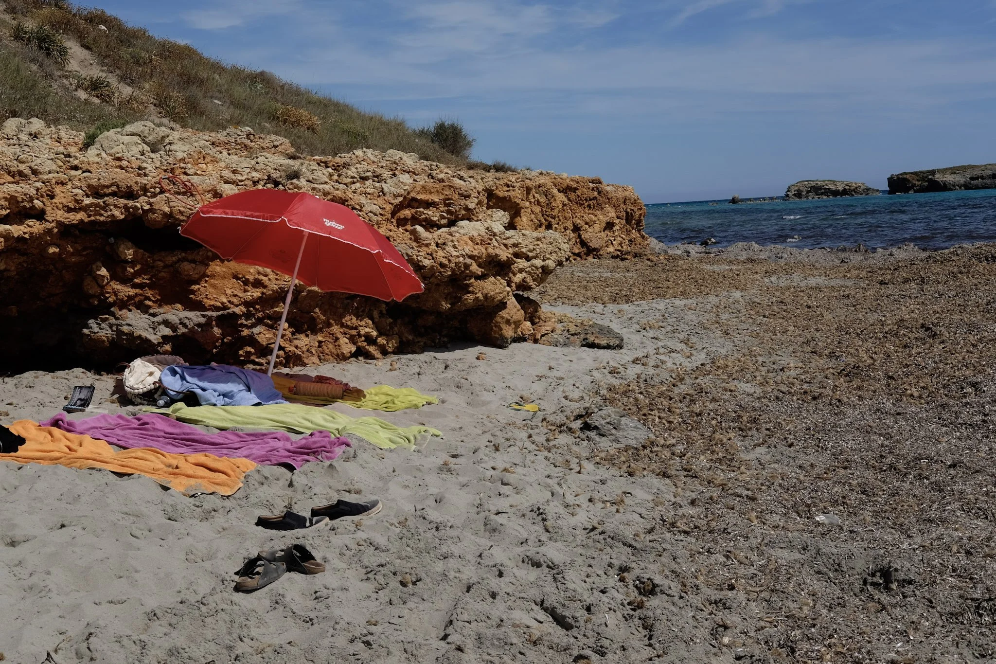 Beach scene with colorful towels, a red umbrella, and various items on the sandy shore. Rocks and hillside with sparse vegetation in the background, and the ocean with small islands or rocks further out under a partly cloudy sky.