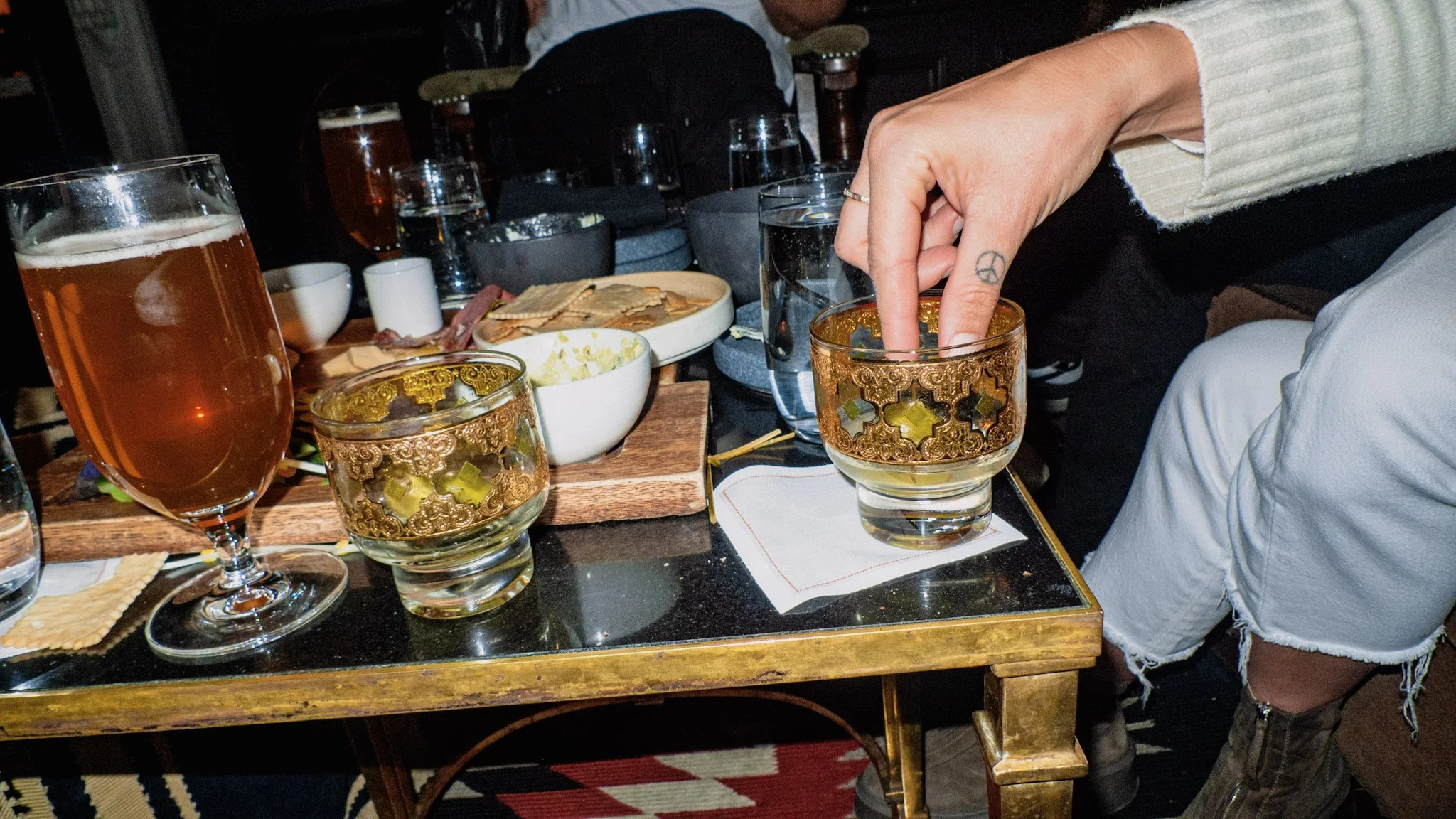 Person's hand with a peace sign tattoo, reaching into an ornate glass on a table at a dinner setting. The table has various dishes, glasses of drinks, and bowls, with other diners visible in the background.