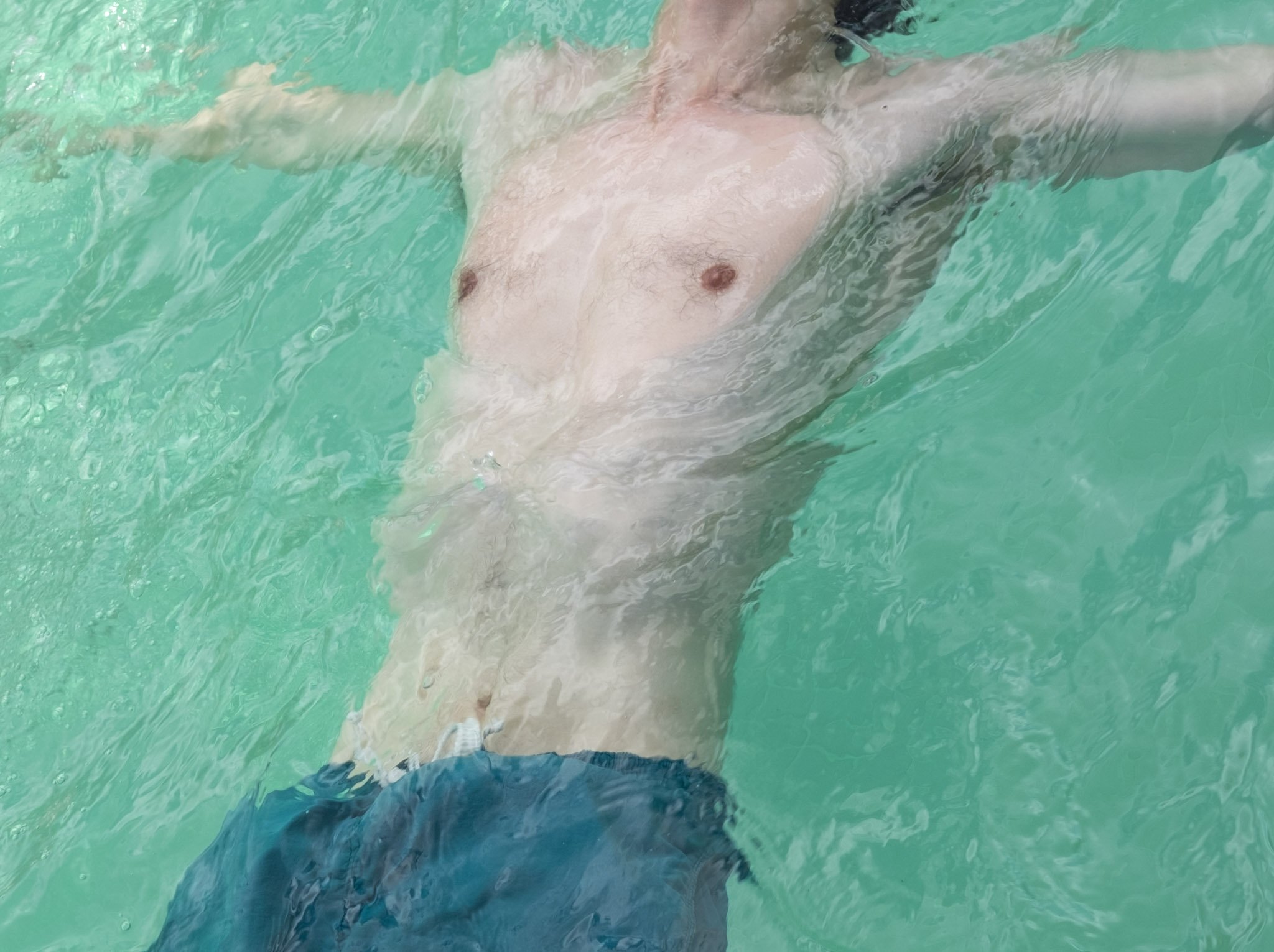Person in swimming pool, lying on pool float, wearing swim trunks and shoes.