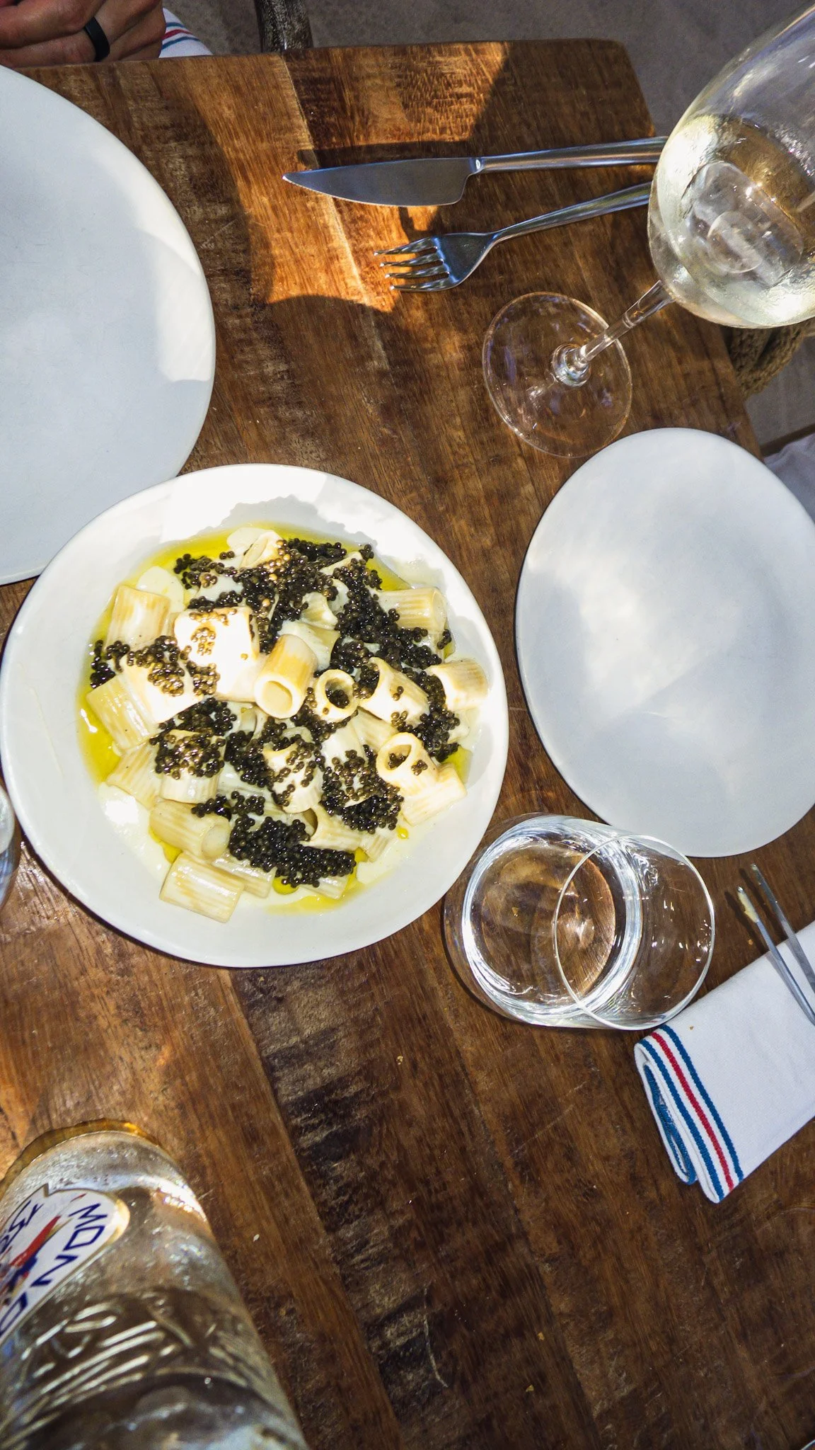 A white plate of pasta with black caviar, placed on a wooden table set for a meal with empty white plates, wine glasses, and water bottles visible.